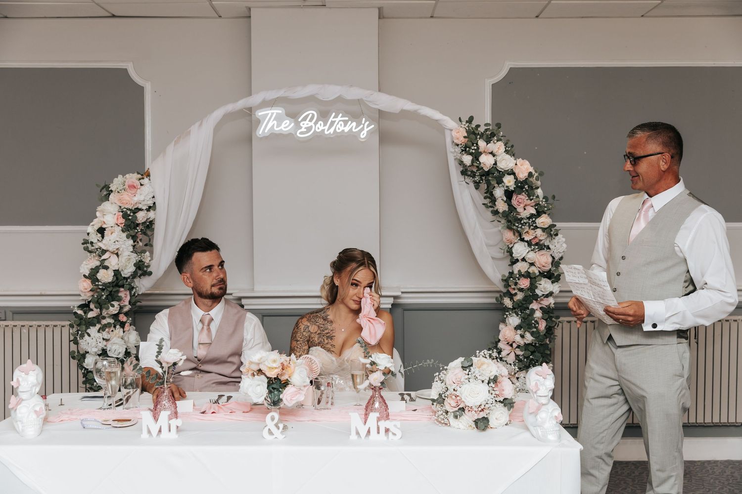 Bride and groom seated at wedding sweetheart table with floral arch and neon sign reading The Boltons.