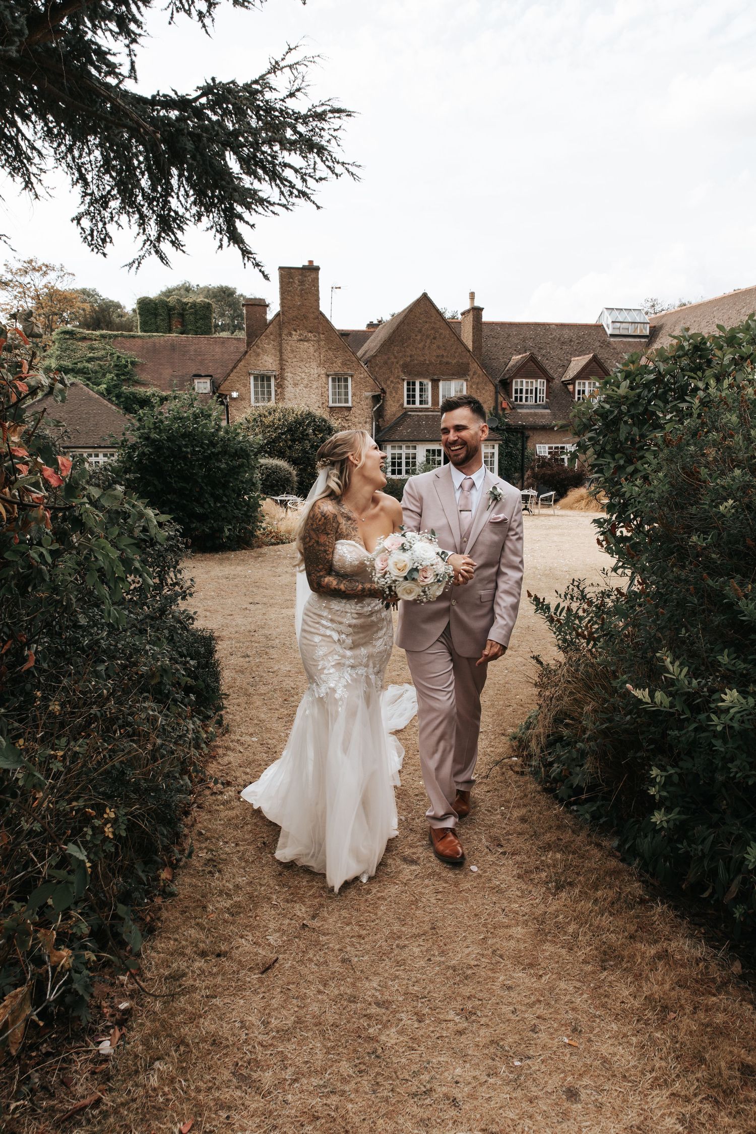 A bride and groom walk hand in hand through a garden path beside a charming brick manor house on their wedding day.