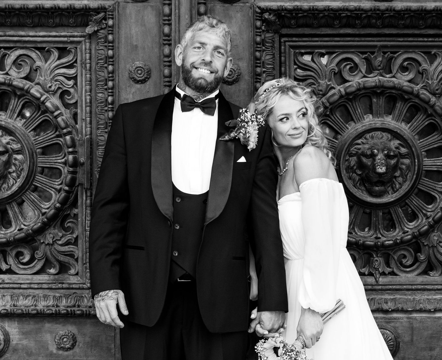 Black and white wedding photo of a smiling couple in formal attire standing before ornate carved wooden doors.