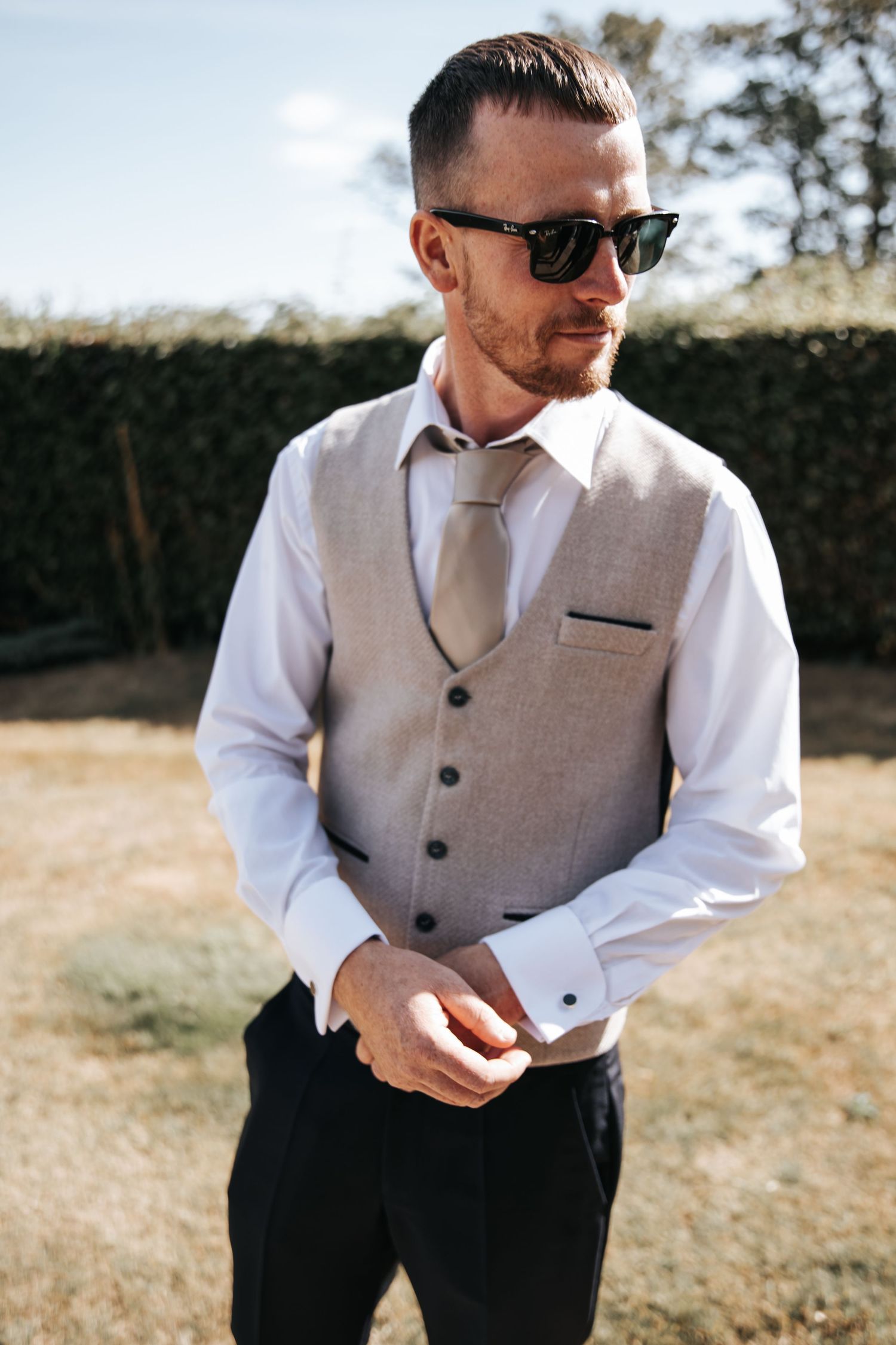 Dapper groom in grey waistcoat, white shirt and tie adjusts cufflinks outdoors on wedding day.