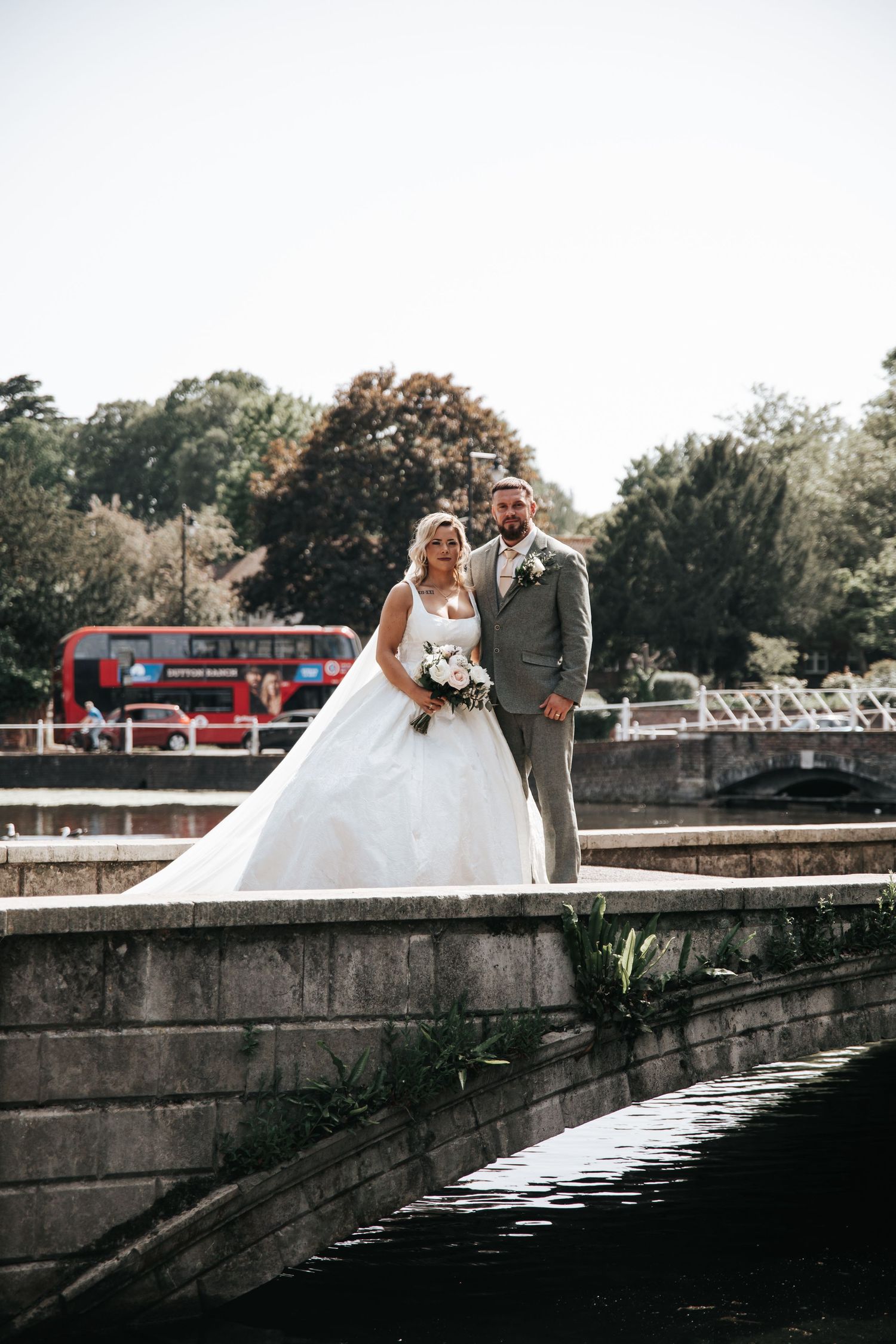 Bride and groom pose on a stone bridge with a red double-decker bus and lush trees in the background.