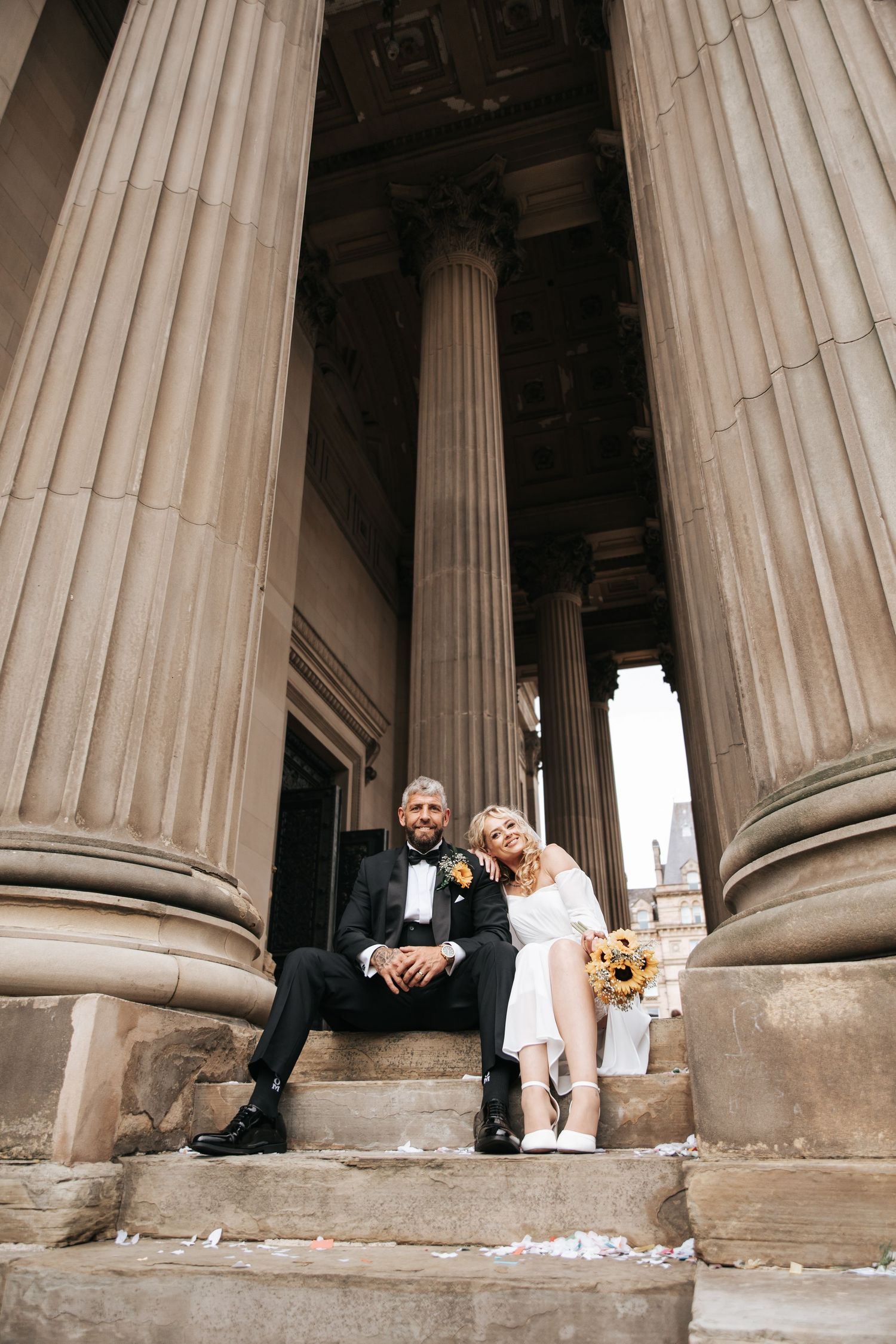 Bride and groom sitting on stone steps between tall classical columns at their wedding venue.