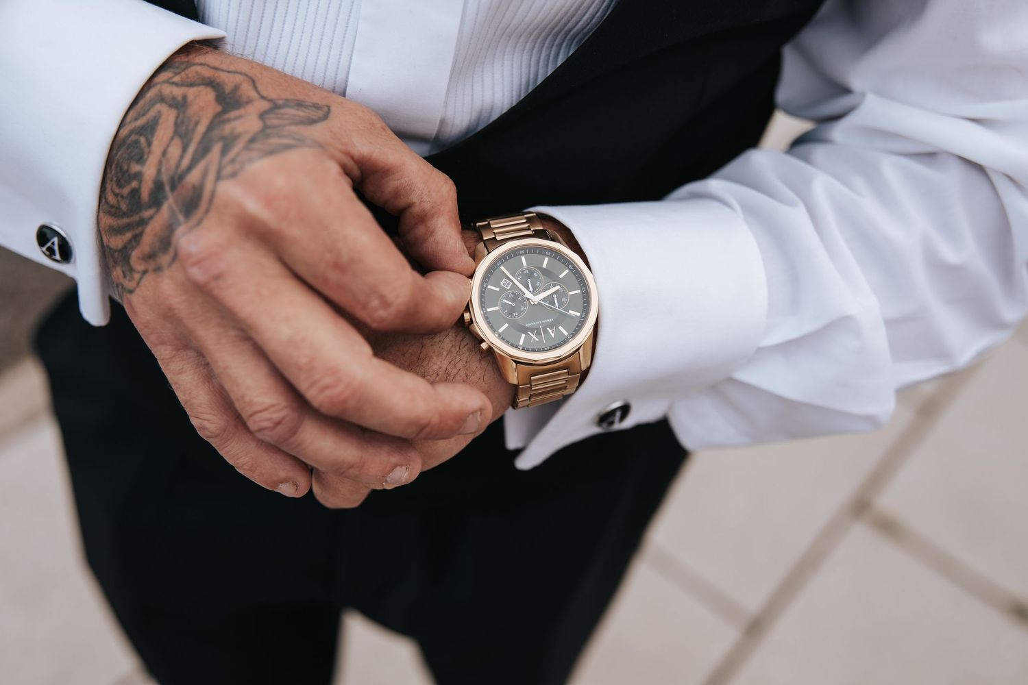 Tattooed man in white dress shirt adjusting gold luxury watch on wrist, elegant formal attire.