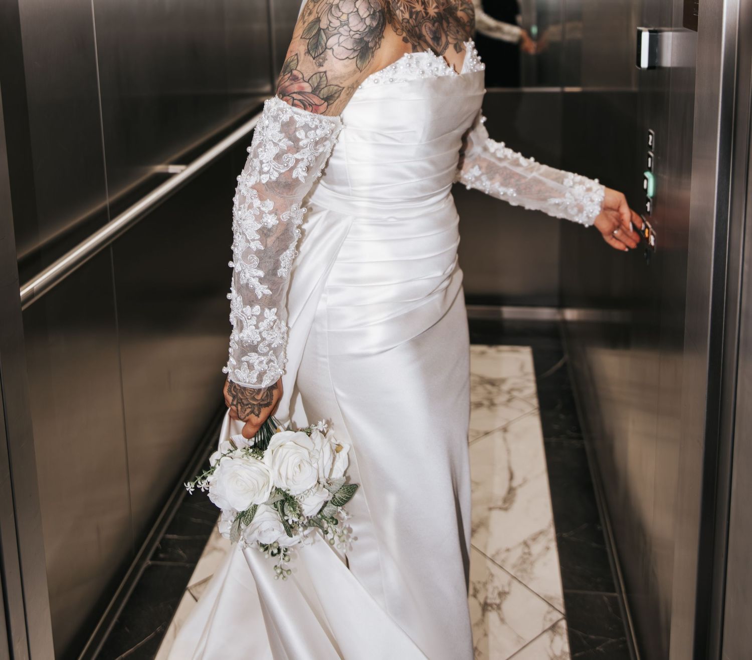 Tattooed bride in white off-shoulder wedding gown holding bouquet in elevator.