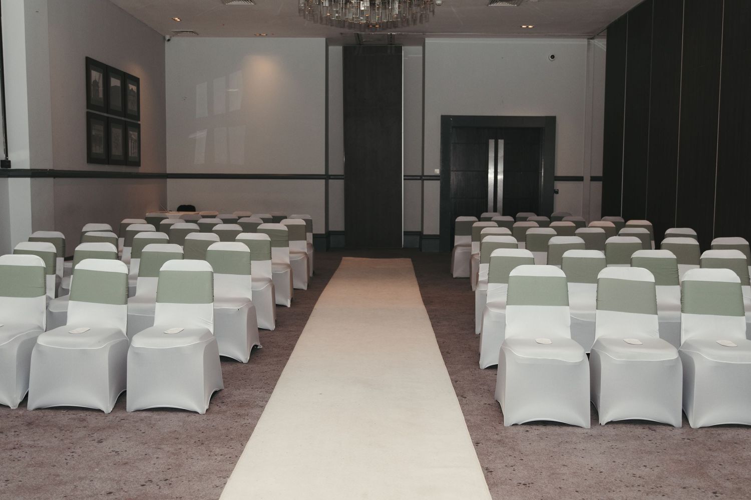 Elegant wedding ceremony venue with white chair covers, sage green sashes, and a white aisle runner in a modern ballroom.