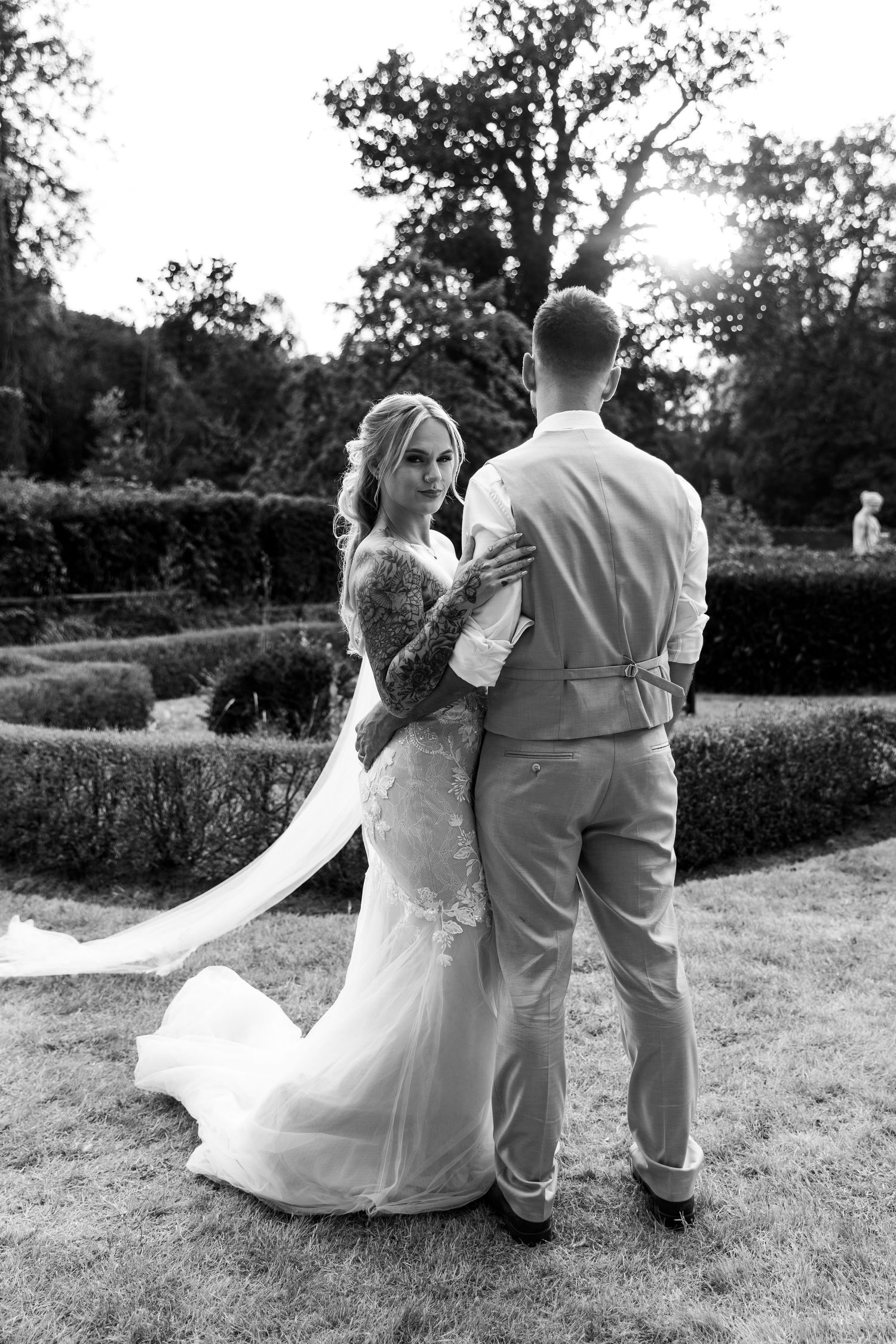 Black and white photo of bride in lace gown embracing groom in formal garden at sunset.