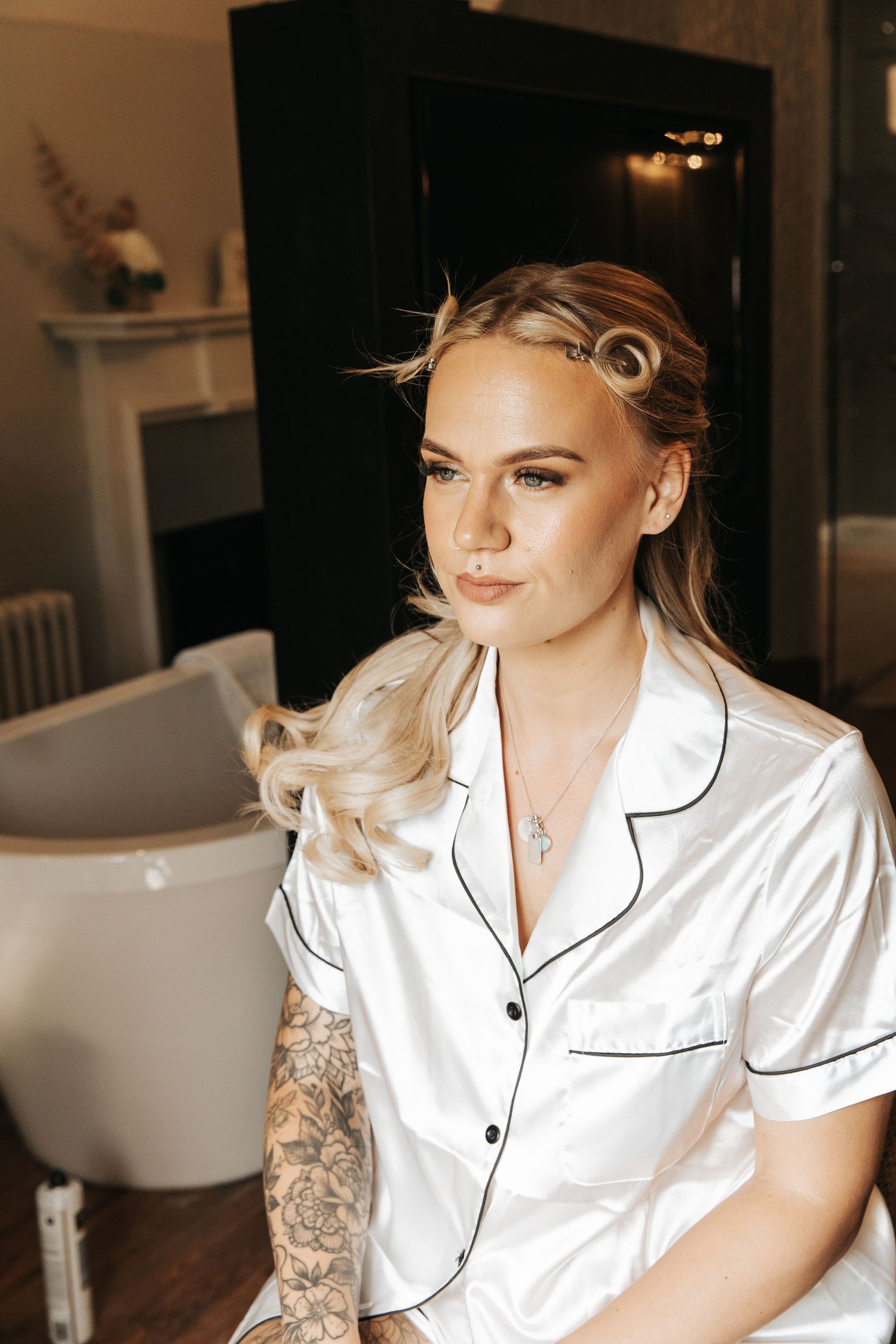 Blonde woman in white satin pajamas with tattoos and a cross necklace sits in a salon getting bridal hair and makeup done.