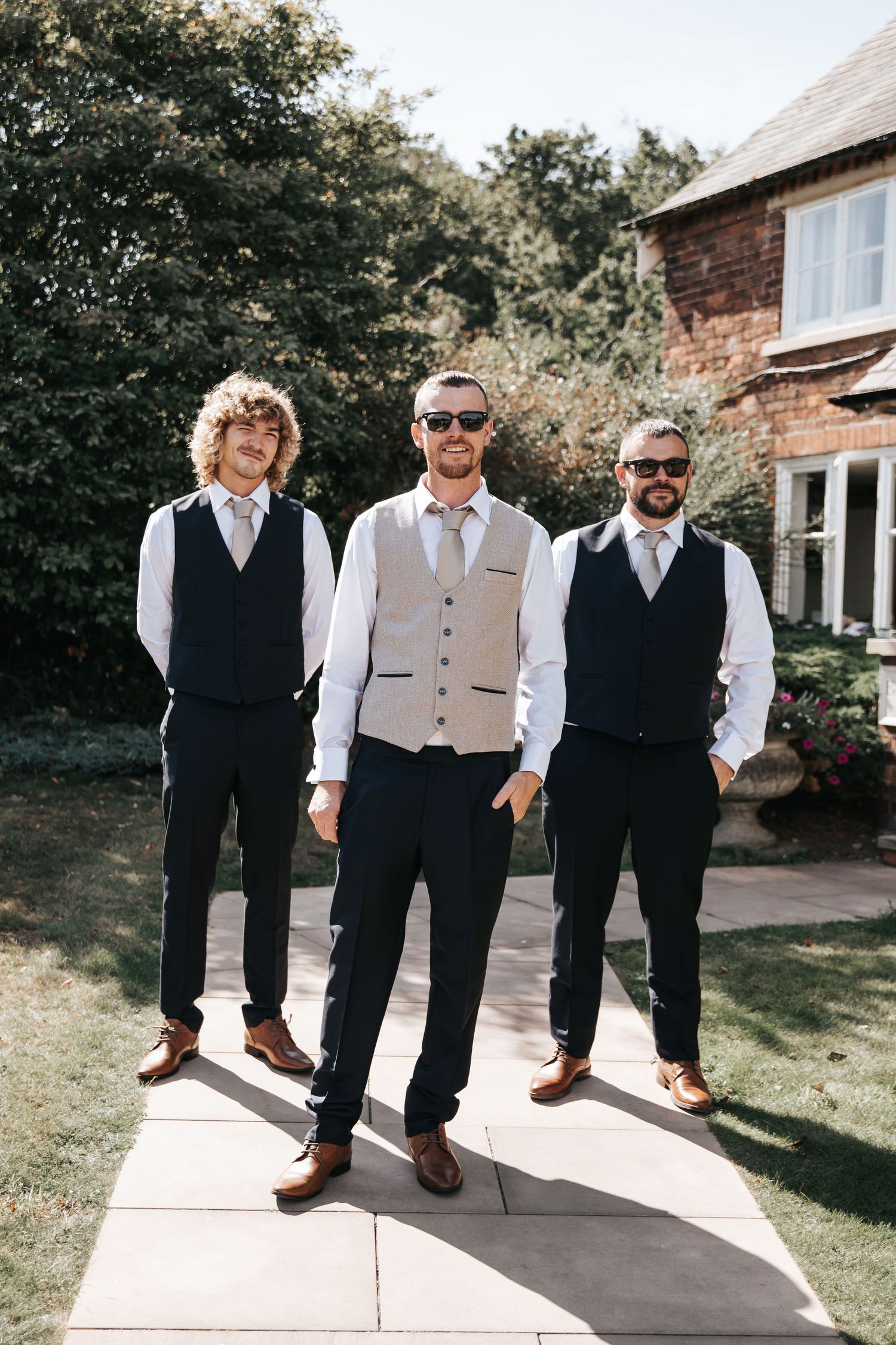 Three groomsmen in navy trousers, waistcoats and sunglasses pose outdoors on a sunny wedding day.