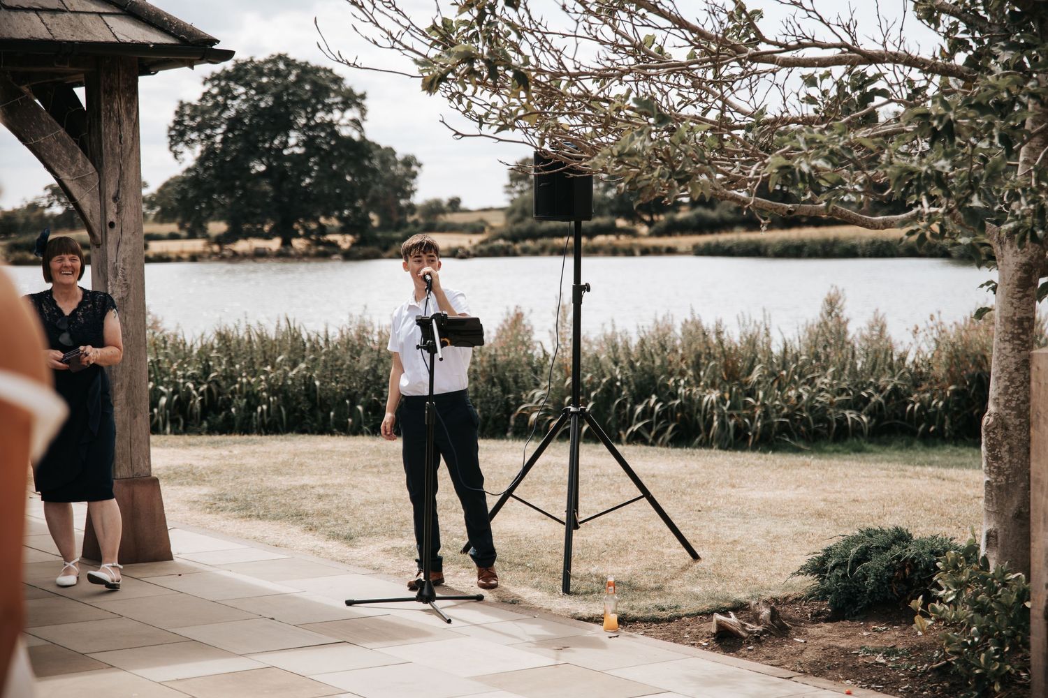 Singer performing outdoors at a lakeside wedding venue with a microphone stand on a sunny day.