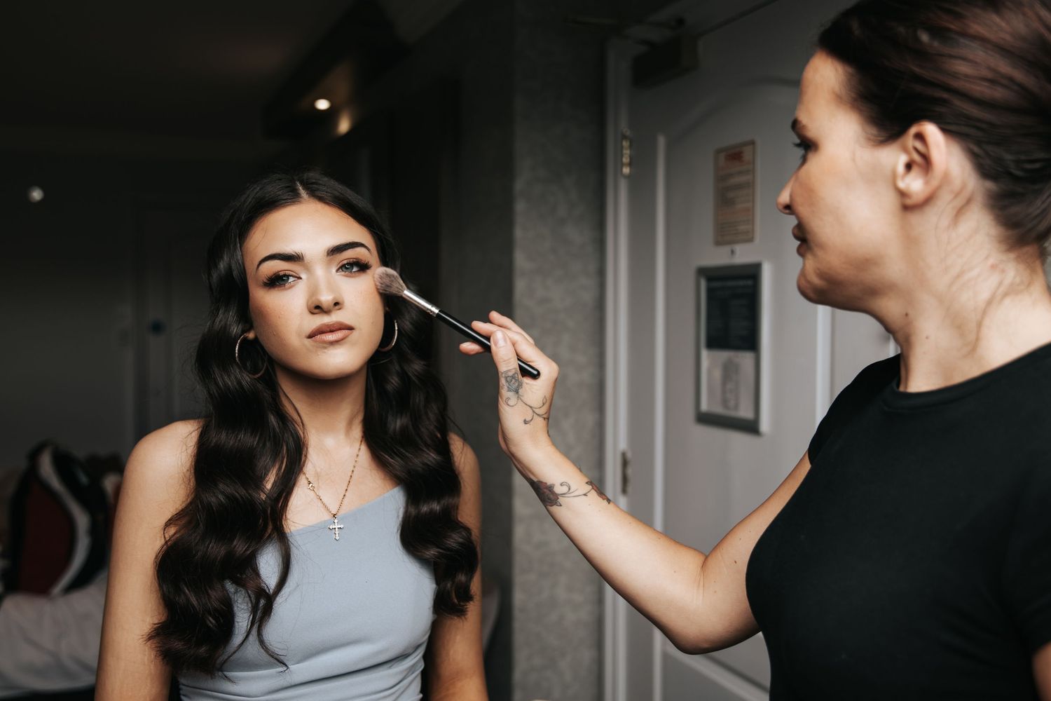 A makeup artist applying foundation to a young woman with long dark wavy hair in a gray dress.