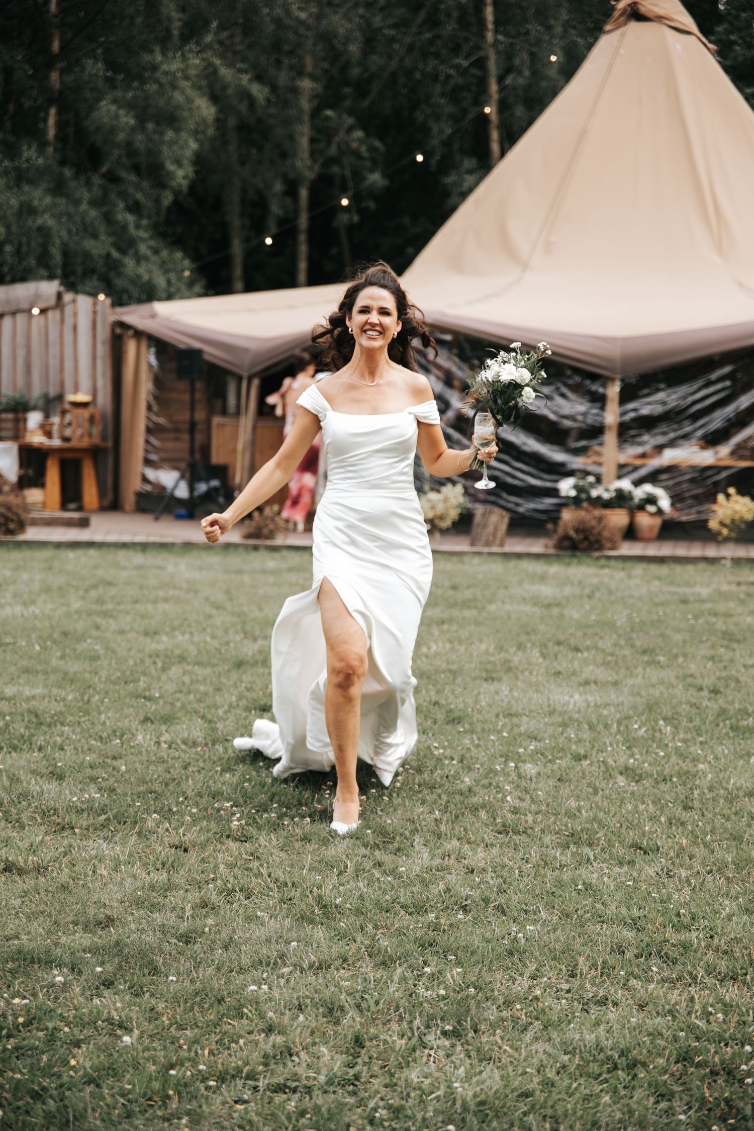 Joyful bride in white off-shoulder gown running on green grass holding bouquet at outdoor wedding venue.