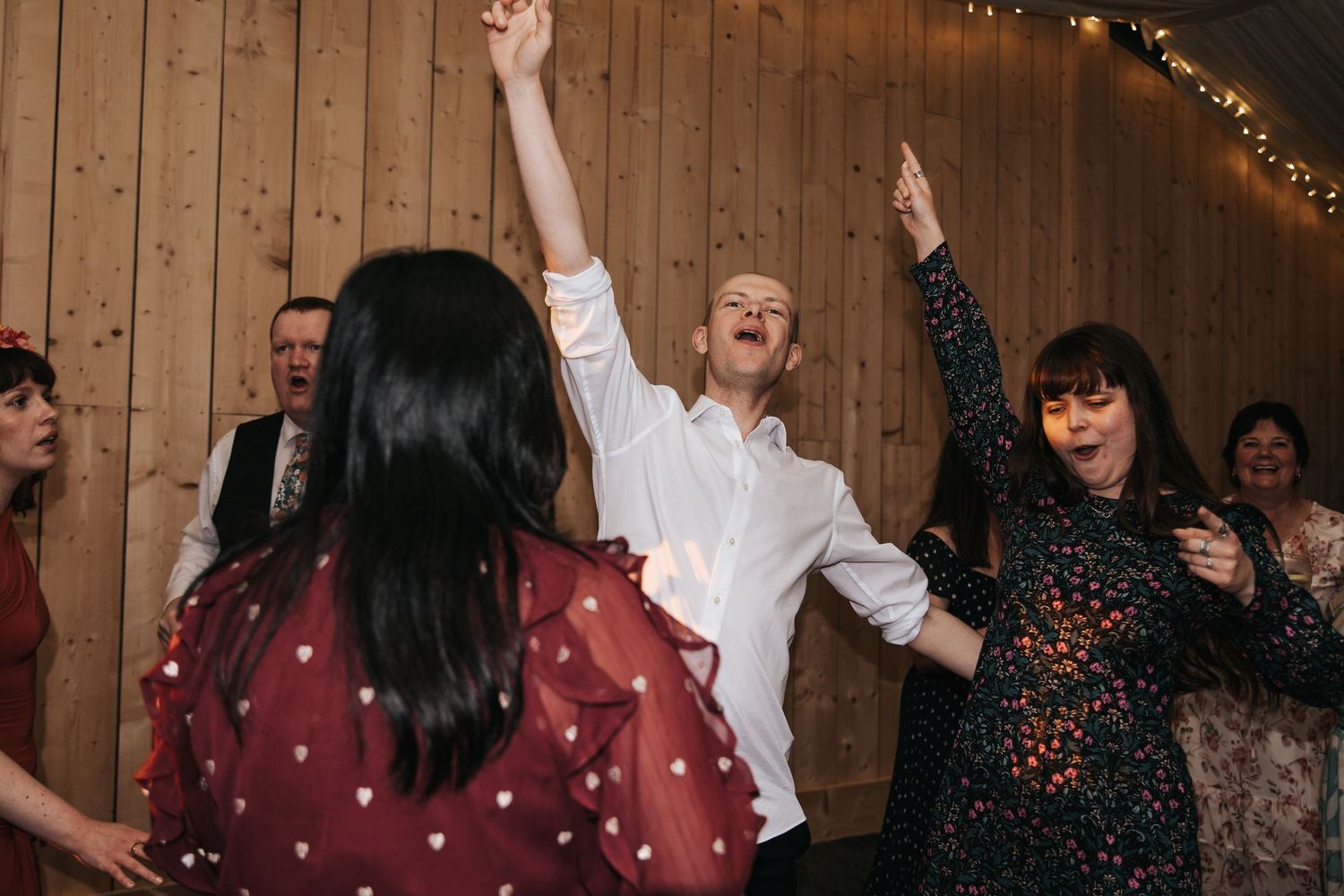 Guests dancing joyfully at a rustic barn wedding reception with fairy lights and wooden walls.