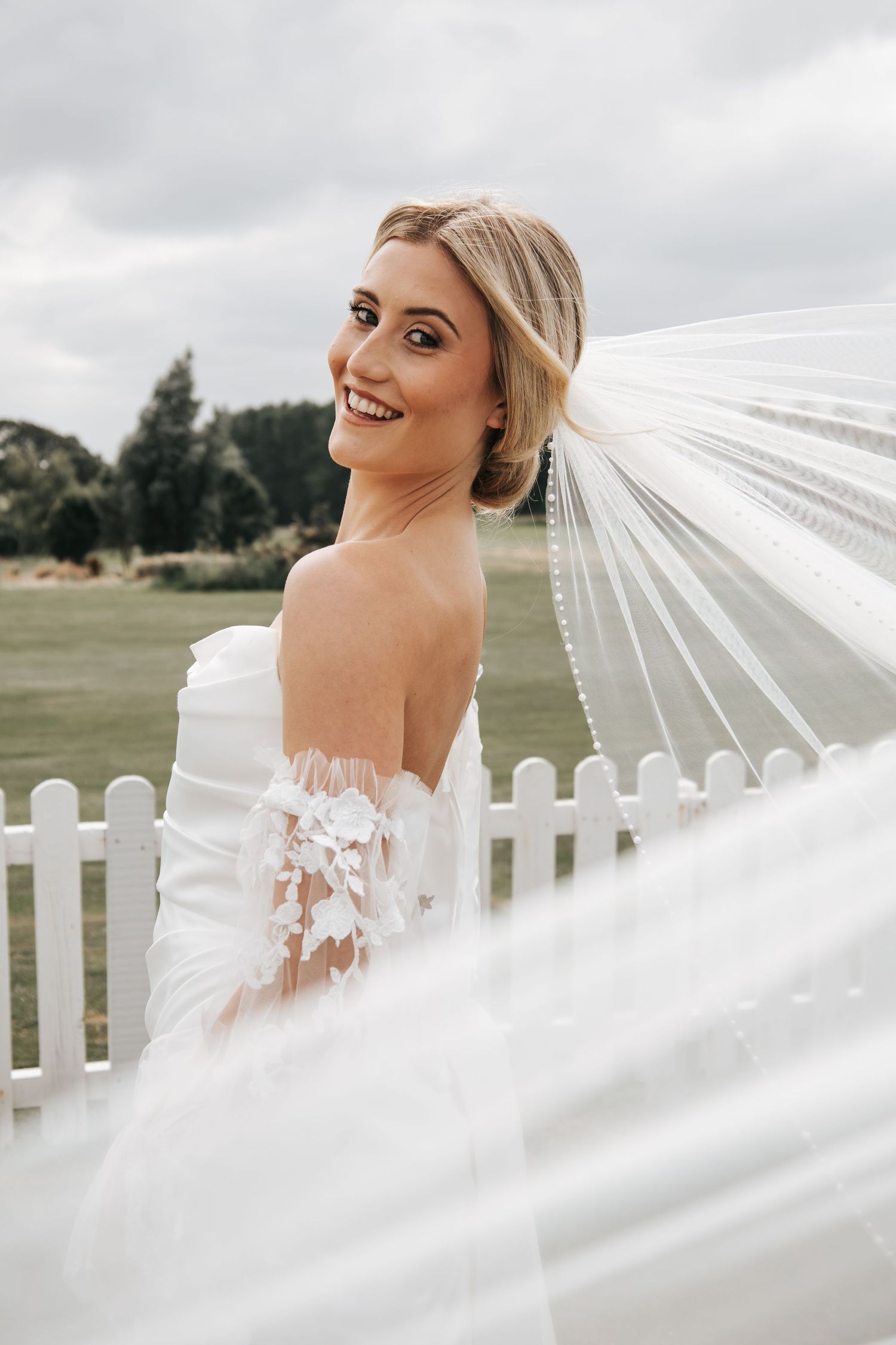 A flowing white wedding veil billows dramatically against a cloudy sky and white picket fence.