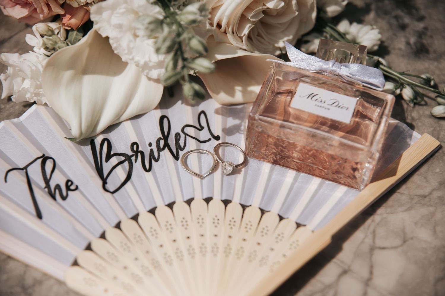 A bridal fan reading The Bride with wedding rings and a Miss Dior perfume box on a marble surface.
