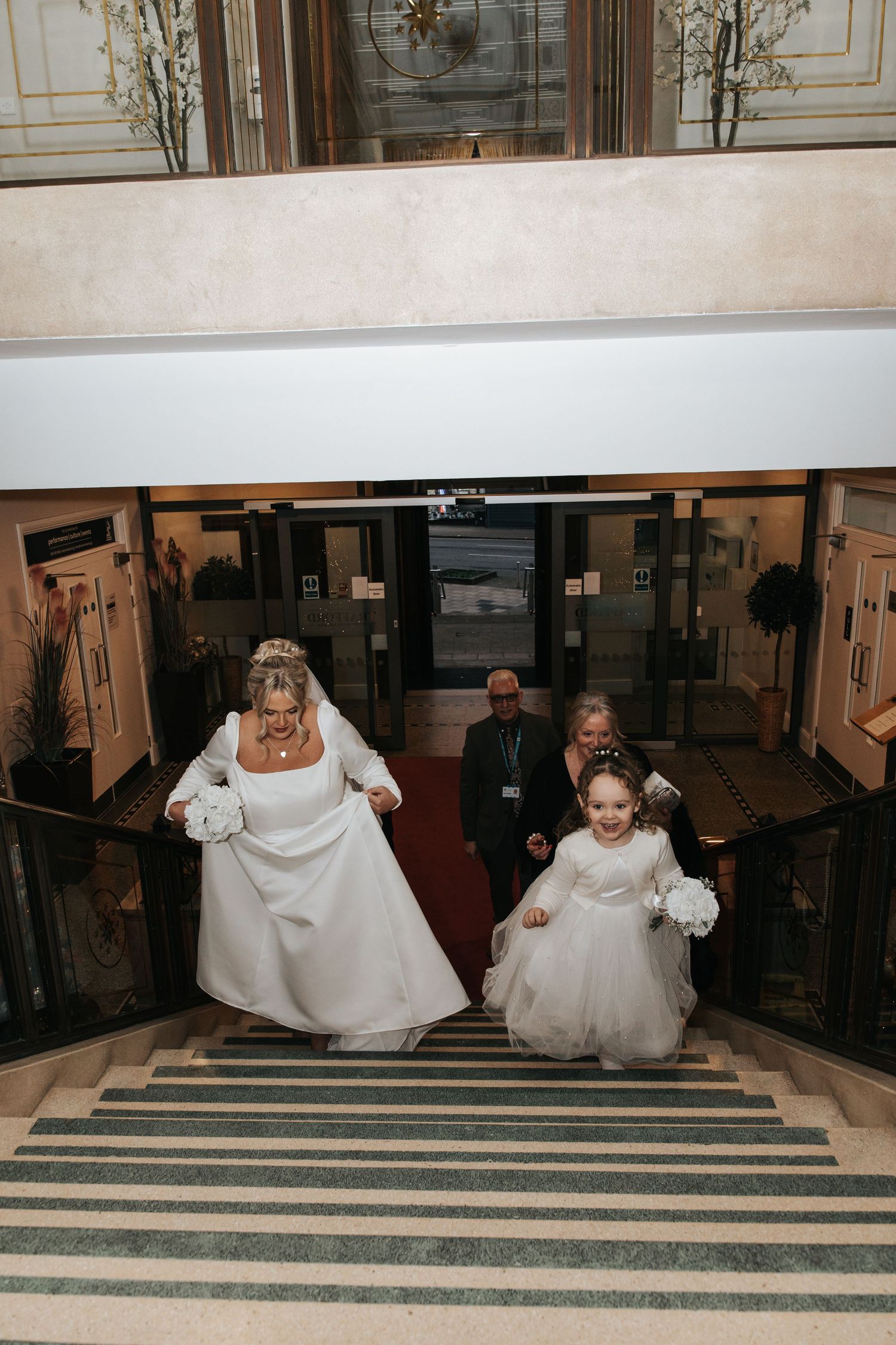 A bride and flower girl descend a grand staircase with red carpet and ornate decor.