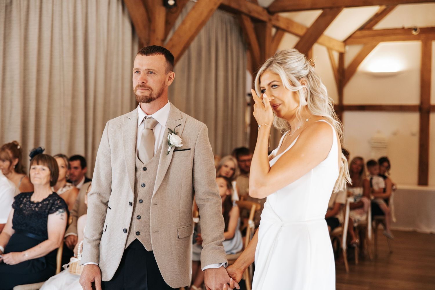 Emotional bride wiping tears while holding hands with groom in beige suit during wedding ceremony in rustic barn venue.