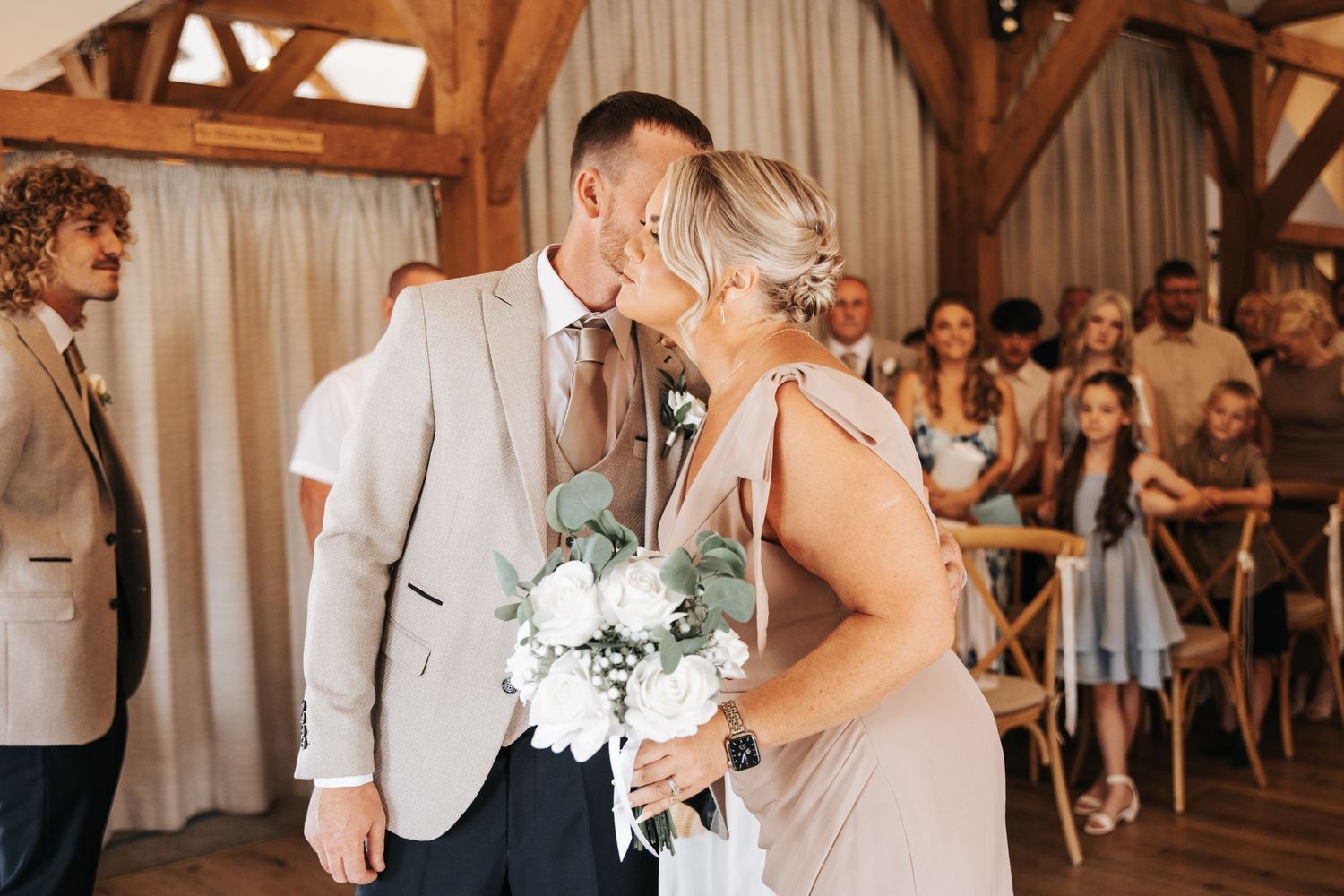 Bride and groom share a first kiss at wedding ceremony in rustic barn venue with guests watching.