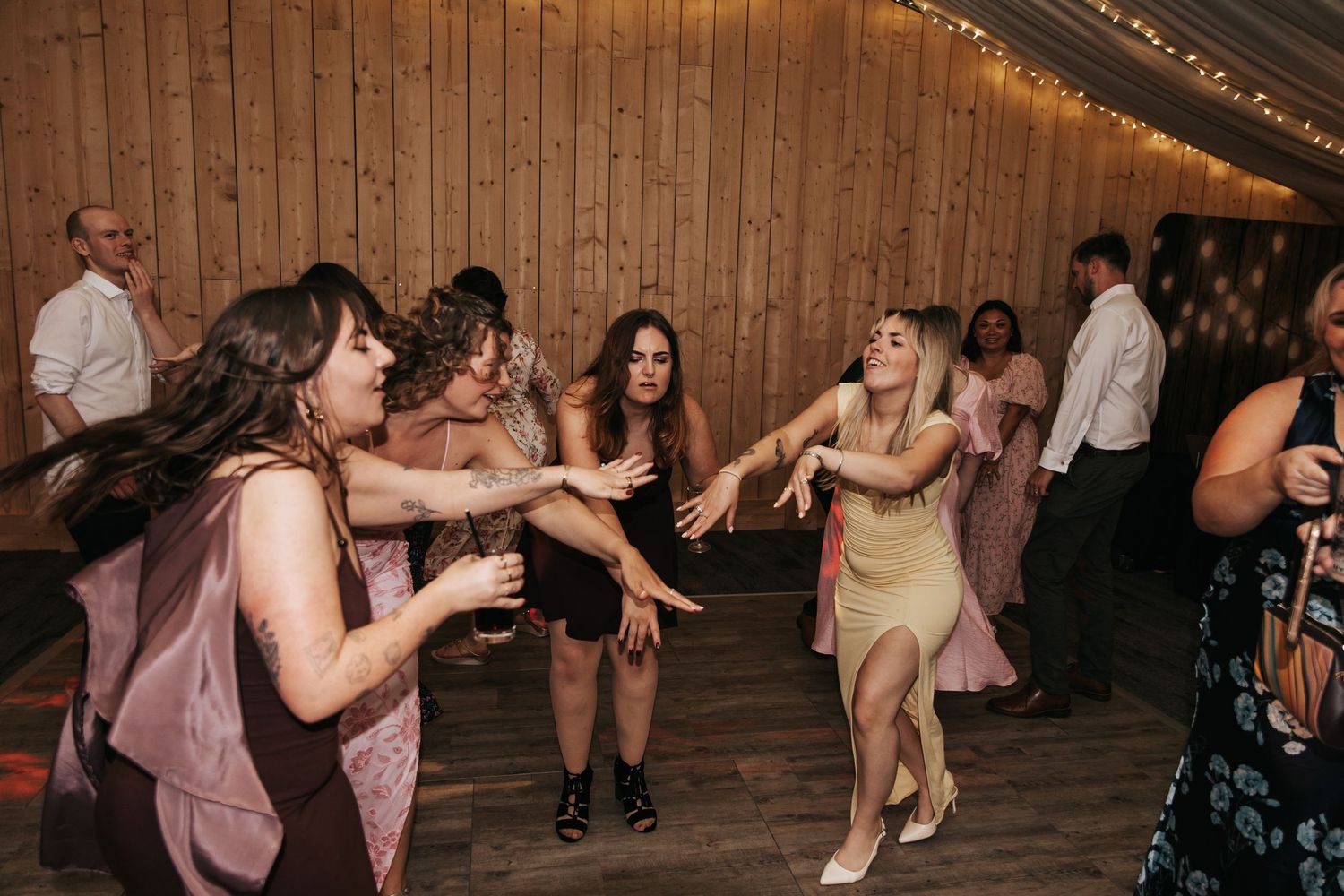 Women dancing energetically at a wedding reception in a rustic venue with warm string lights.