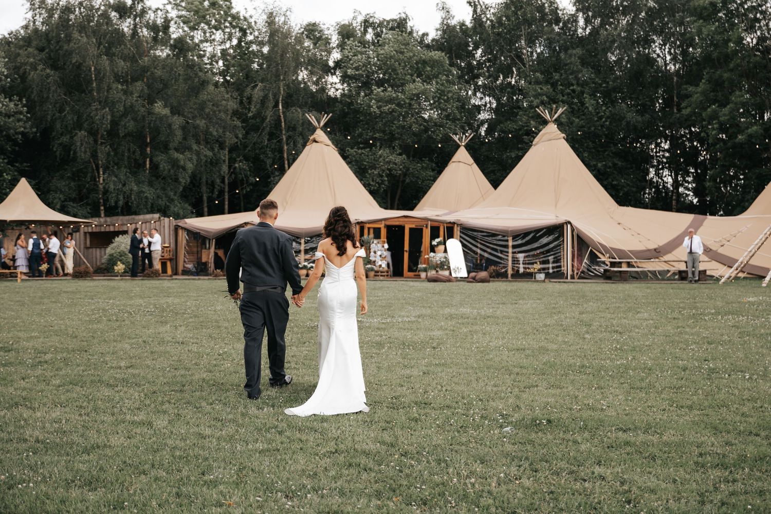 Bride and groom walking hand in hand toward elegant tipi wedding venue on lush green lawn.