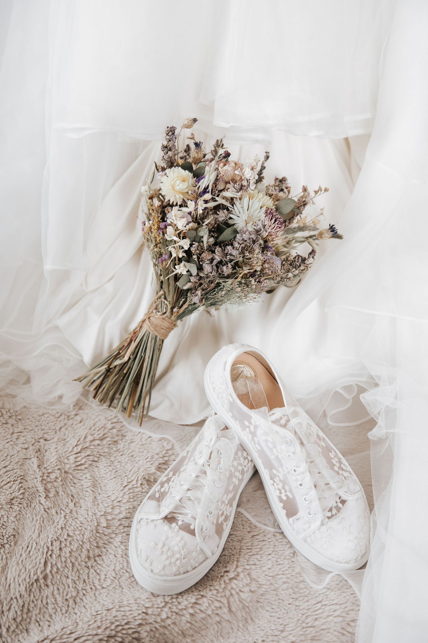 White lace bridal sneakers beside a dried flower bouquet on a fluffy rug with a wedding dress in the background.