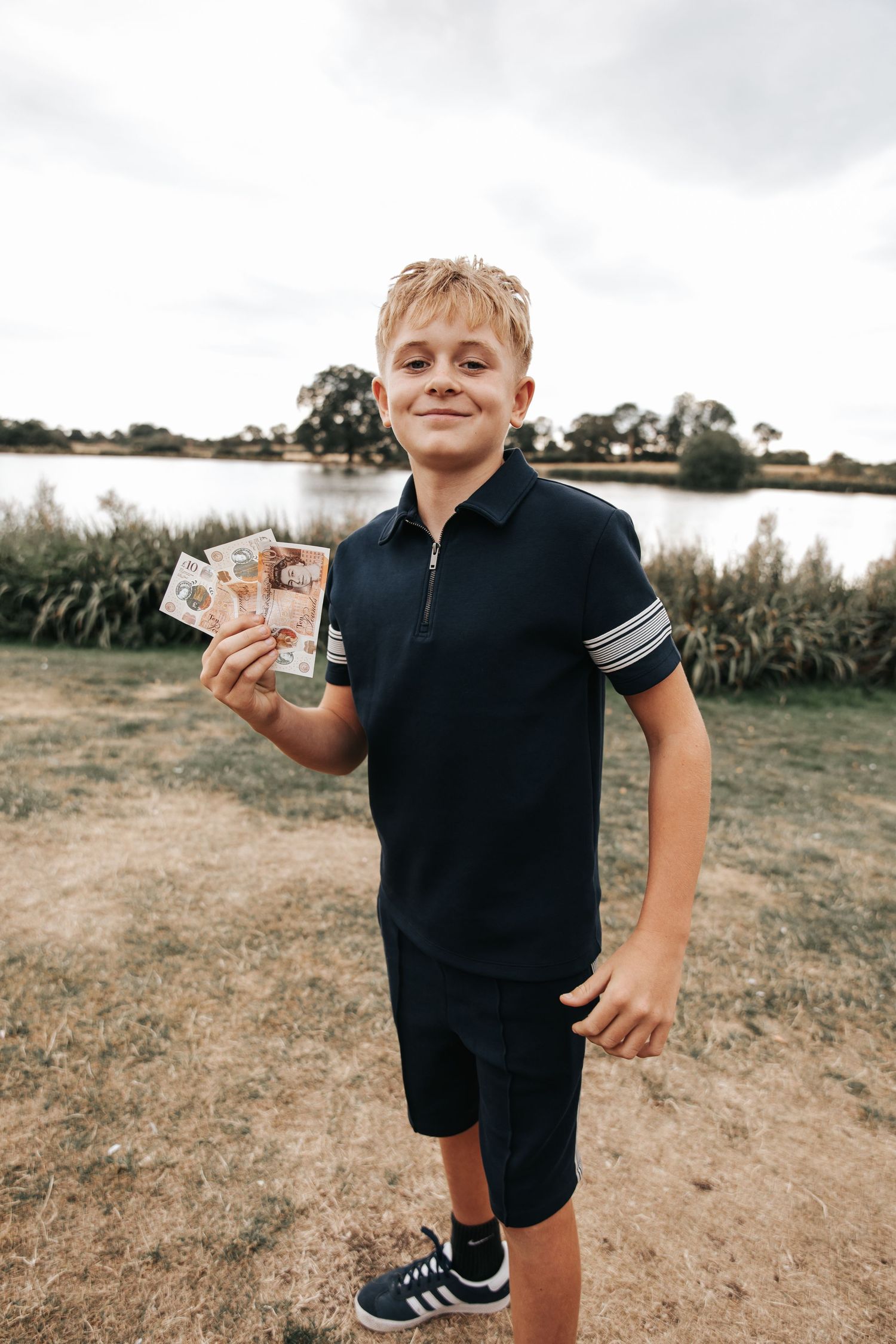 Smiling boy in navy polo shirt holds British pound notes near scenic lake.