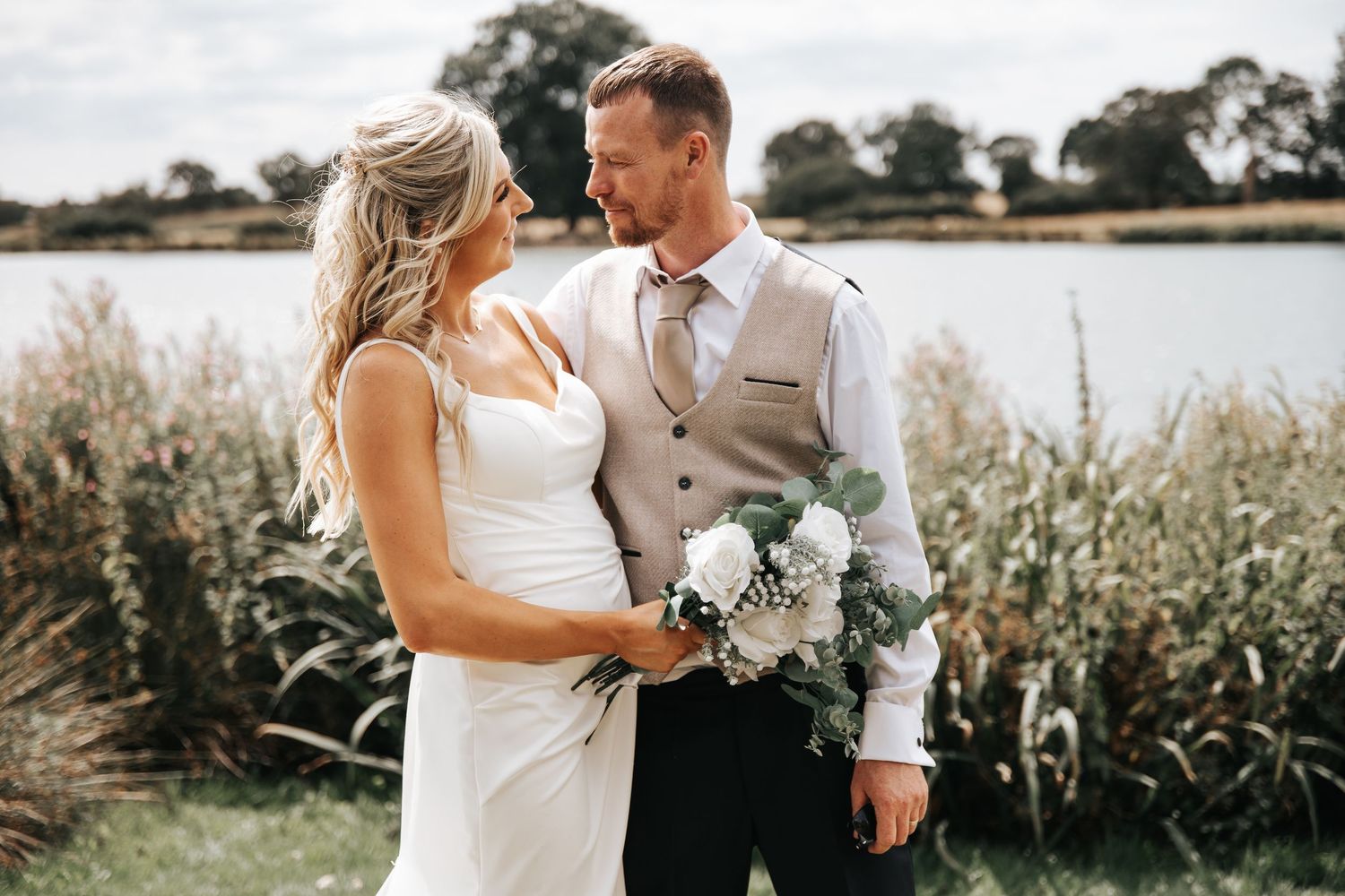 Bride and groom smiling at each other outdoors near a lake, holding a white bouquet, dressed in wedding attire.