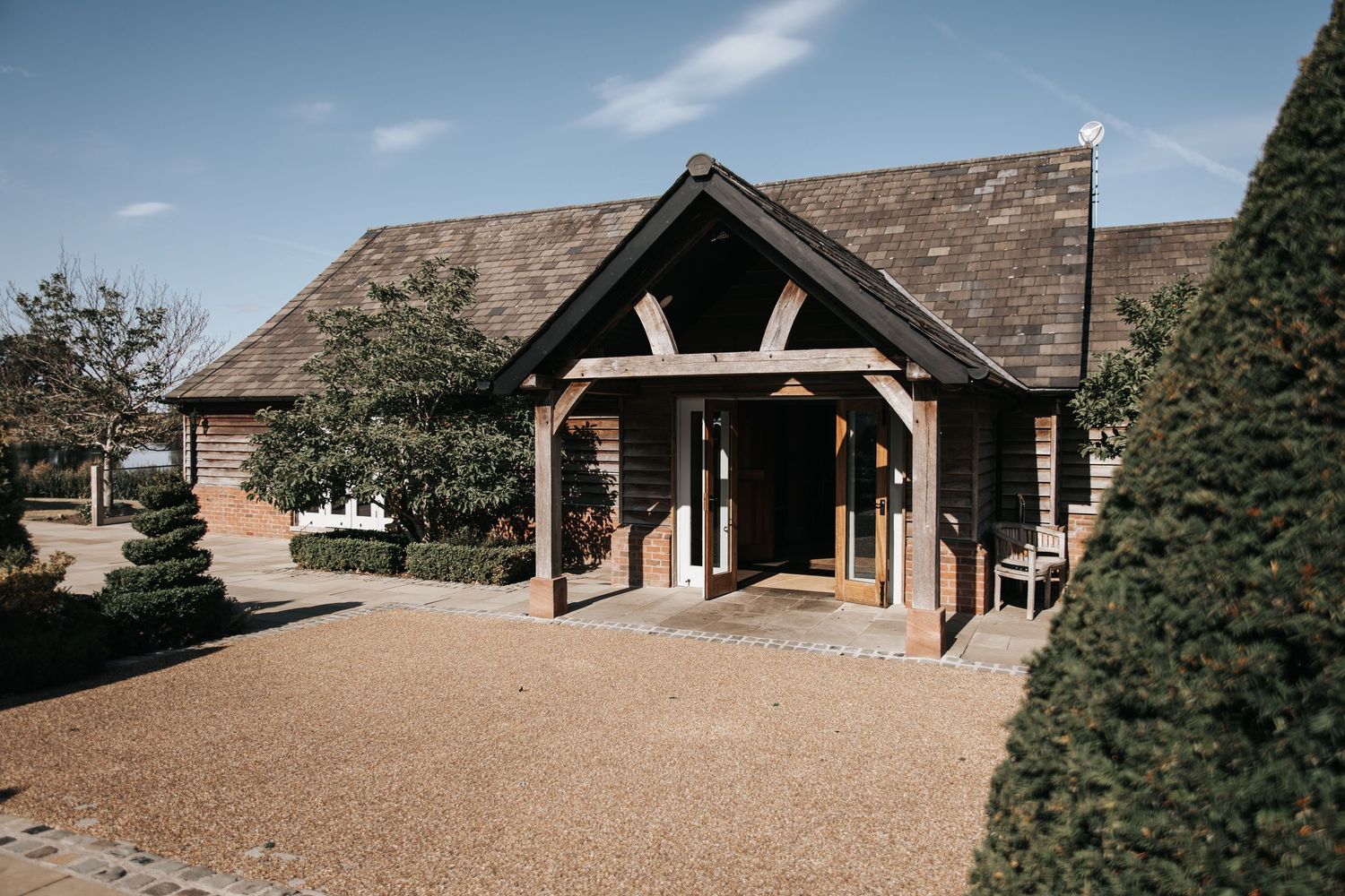 Charming oak-framed barn venue with gravel driveway, manicured hedges, and slate roof under a blue sky.