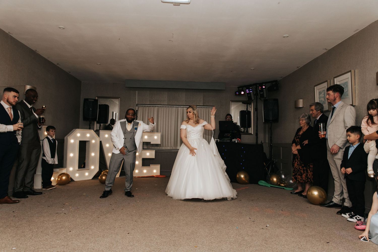 Wedding guests dance and celebrate on a reception dance floor with a DJ setup and decorative lighting.