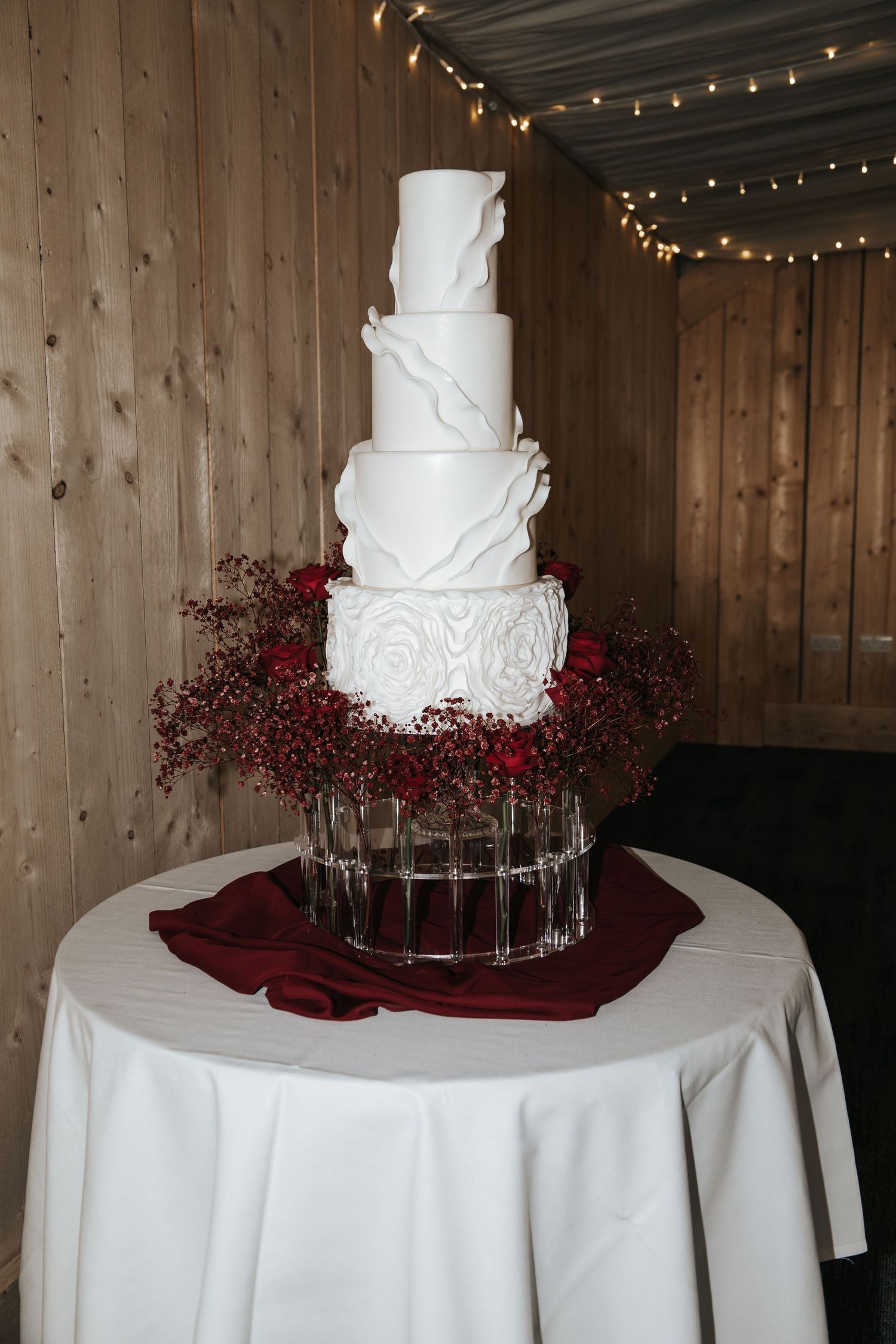 Elegant five-tier white wedding cake decorated with intricate details and red floral accents on a round table.