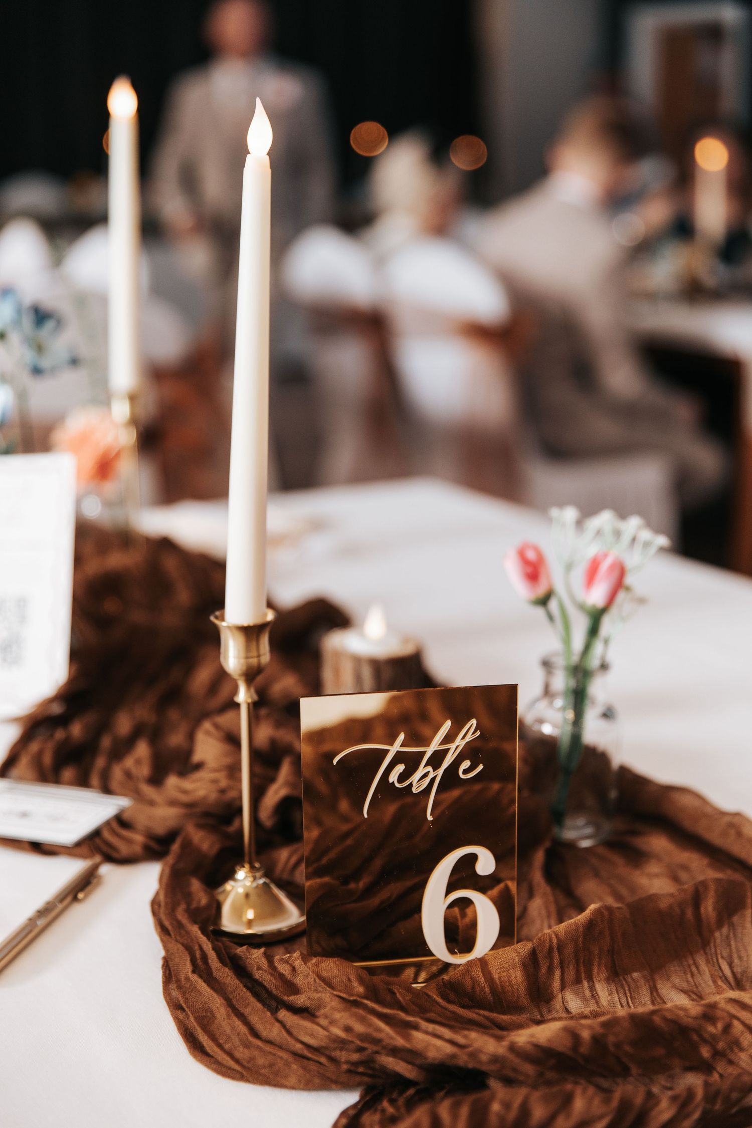 Elegant wedding table setting with gold candlestick, white taper candle, brown linen runner, and Table 6 sign with pink tulips.