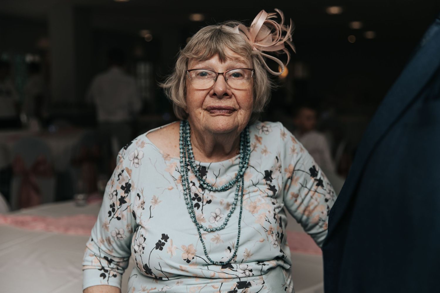 Elderly woman wearing glasses and teal beaded necklace with floral dress and feathered hair accessory at indoor event.