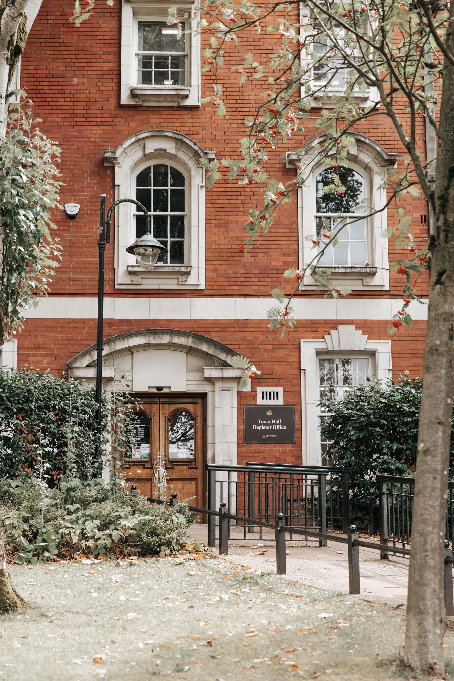 Historic red brick building with arched windows, wooden doors, and a Town Hall Register Office sign, framed by autumn trees.