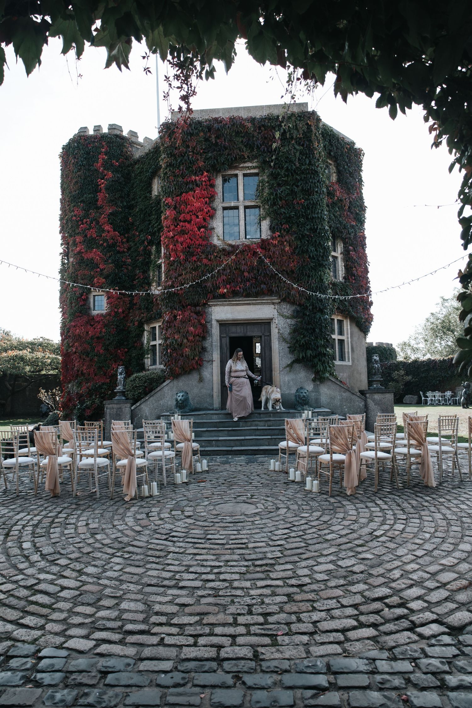 Ivy-covered castle venue with cobblestone courtyard, white chairs arranged for outdoor wedding ceremony, red autumn foliage.