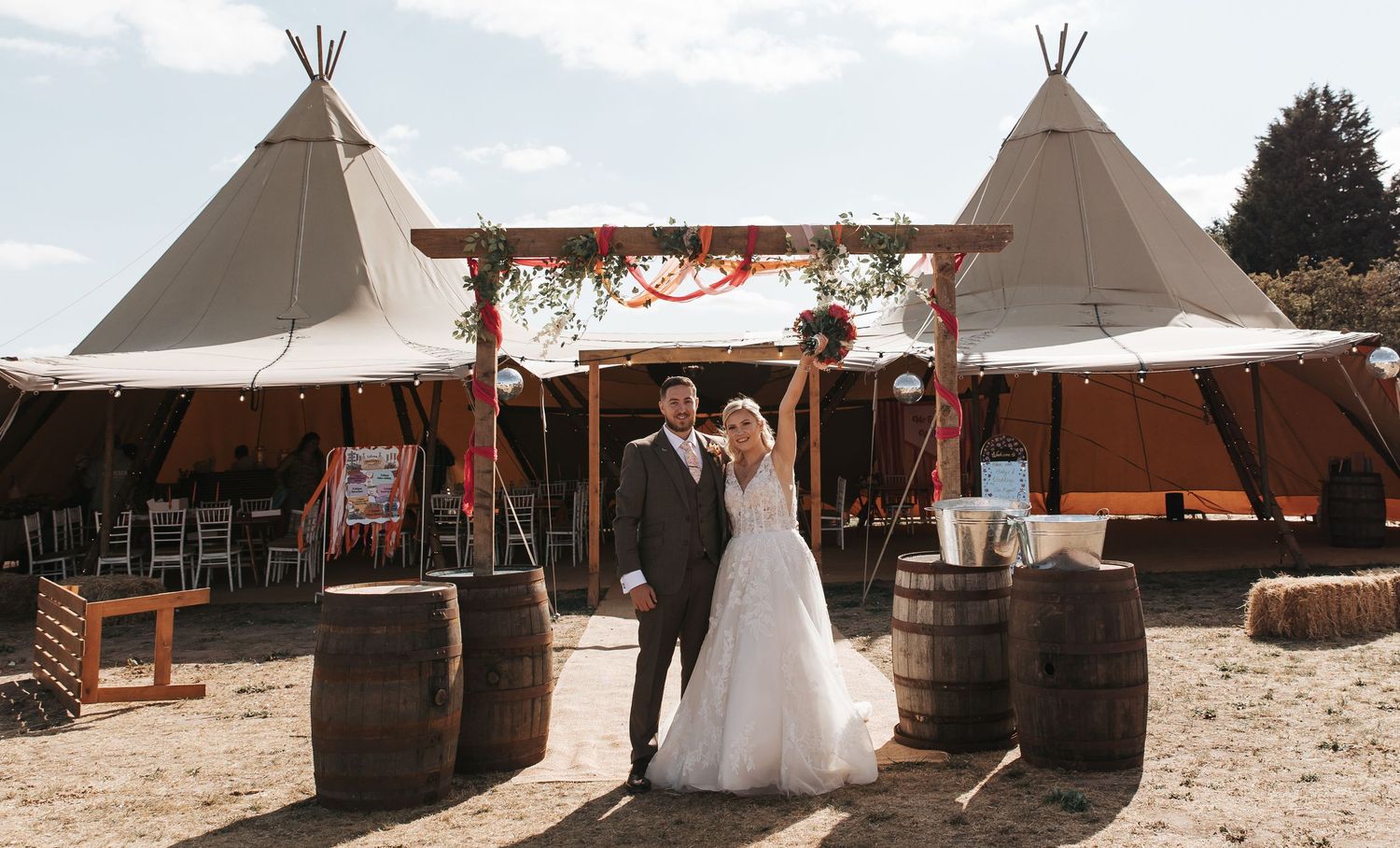 Bride and groom standing together at a rustic tipi wedding venue with wooden barrels and floral decorations.