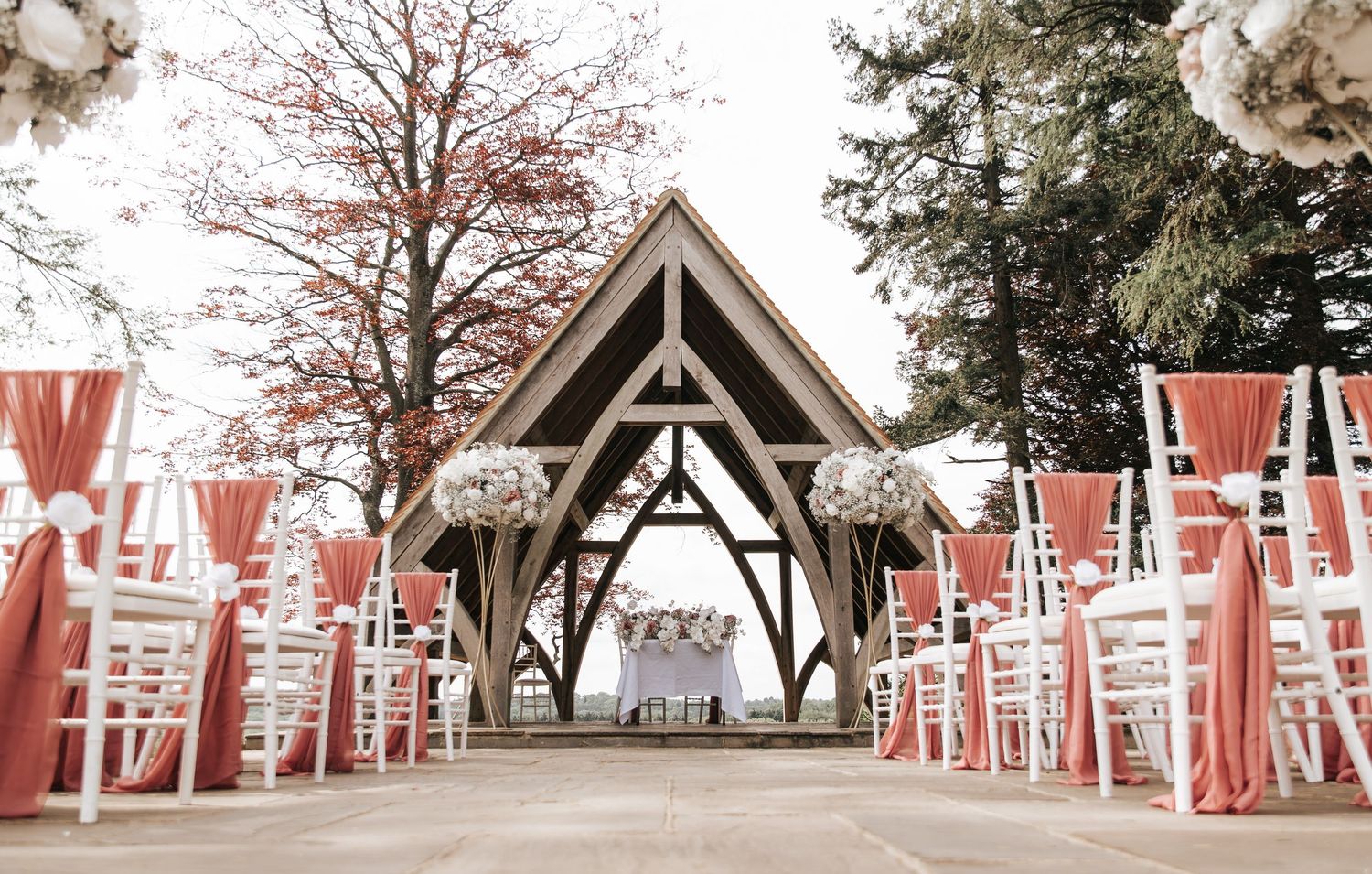 Elegant outdoor wedding ceremony with white chairs, pink draping, and floral arrangements under a wooden arch.