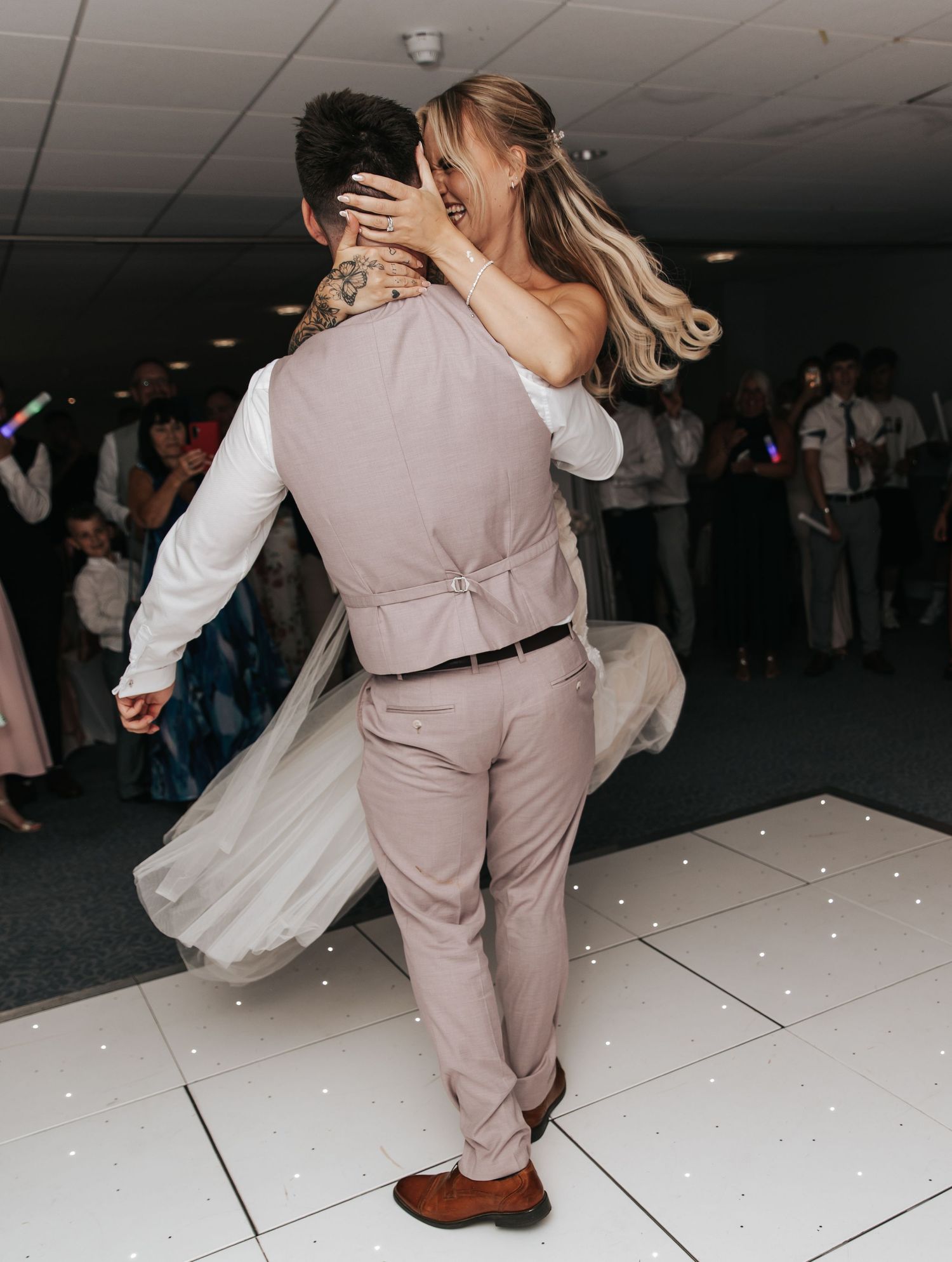 Bride and groom sharing first dance, groom lifting bride in pink wedding dress on white sparkle dance floor.