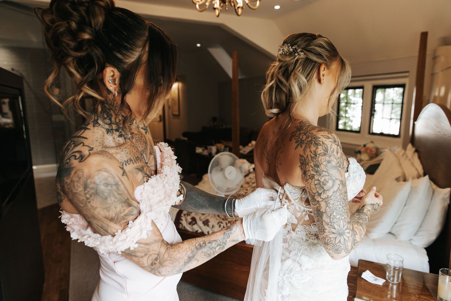 Two tattooed brides in white wedding gowns helping each other dress in an elegant room.