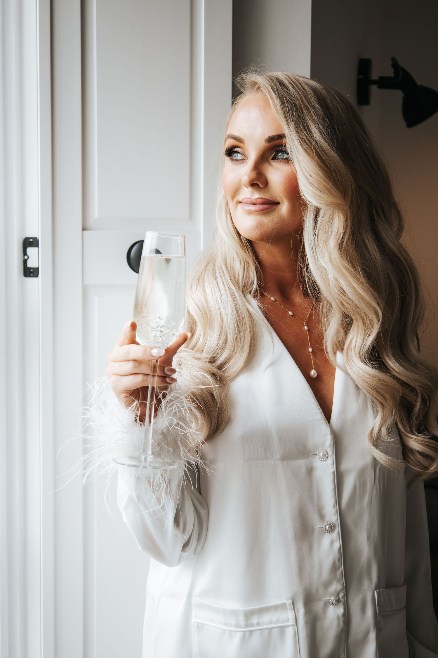 Woman in white robe holds champagne glass while getting ready.