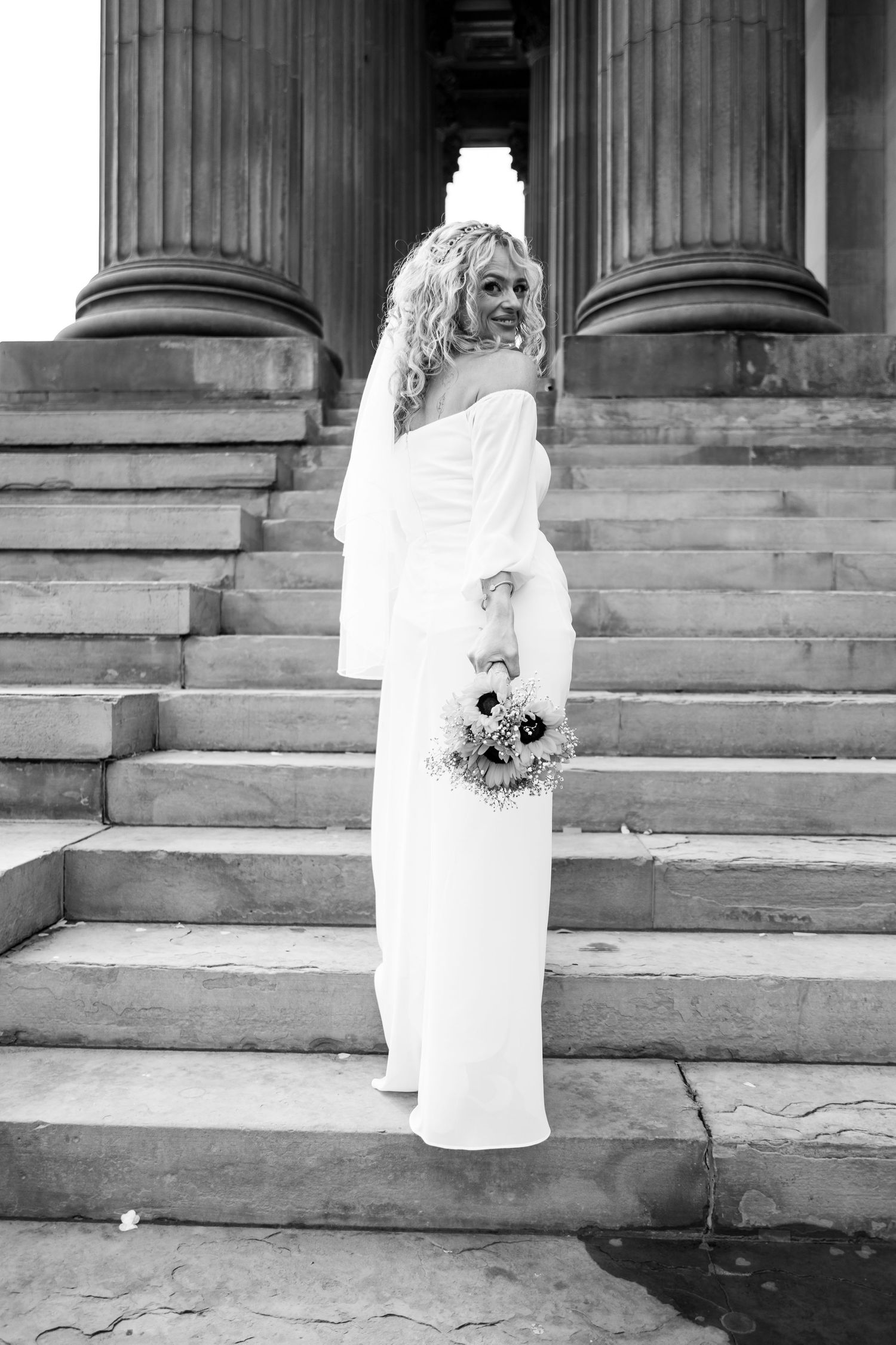 Black and white photo of a bride in a white off-shoulder gown holding a bouquet on stone steps with columns.