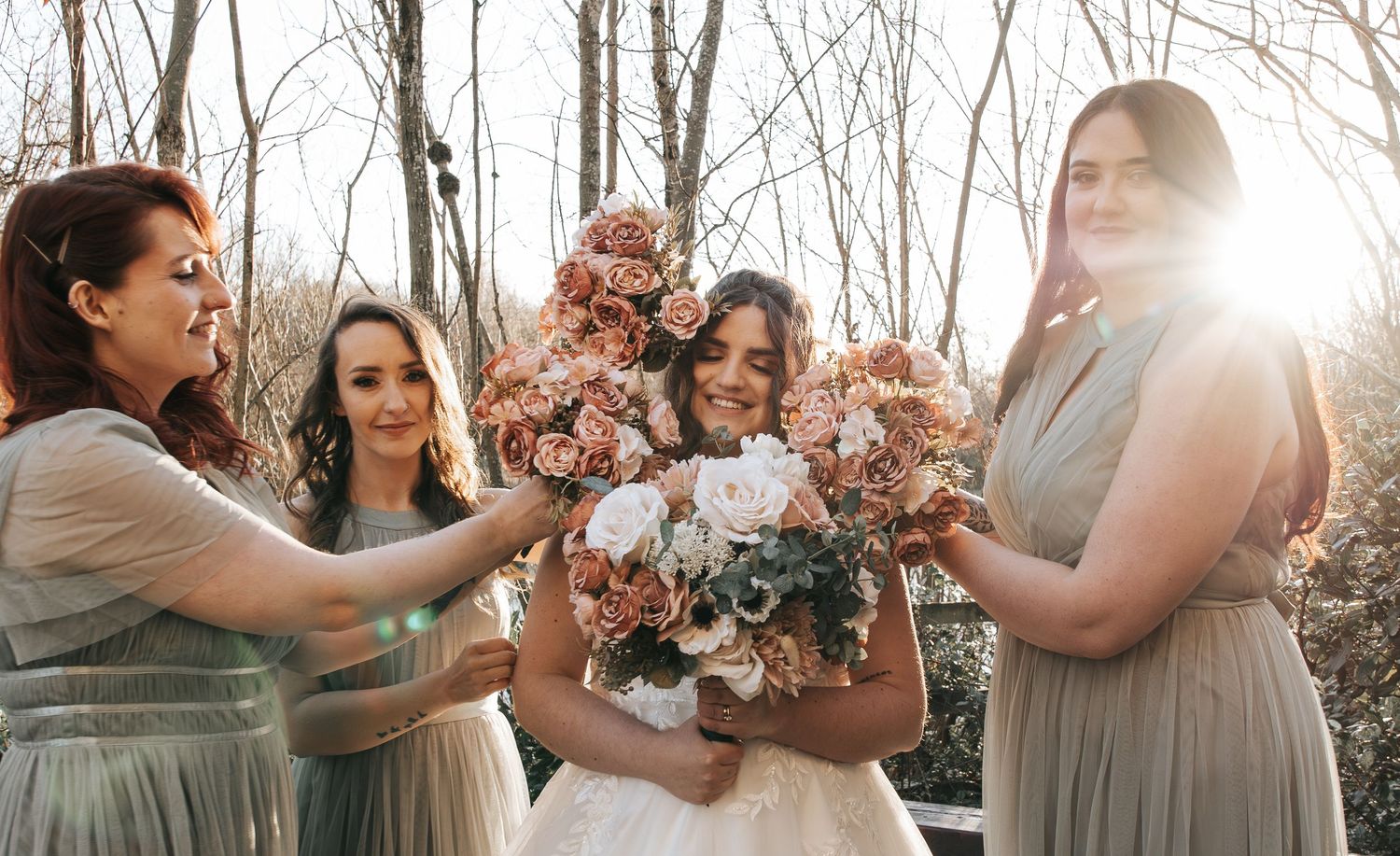 Bridesmaids in grey dresses share joyful moment with bouquets in sunlit forest setting.
