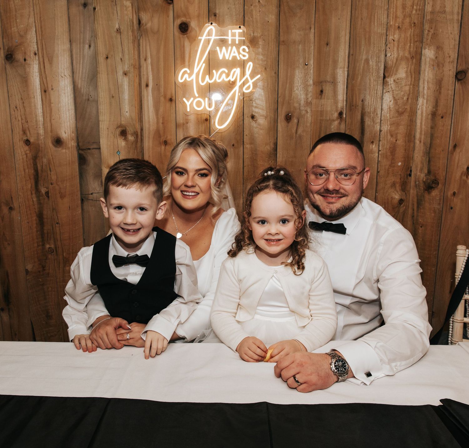 Wedding party poses beneath neon sign against rustic wooden wall during reception celebration.