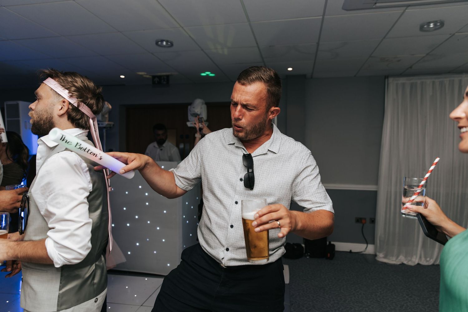 Two men dancing at a wedding reception, one holding an iced drink, in a decorated event venue.