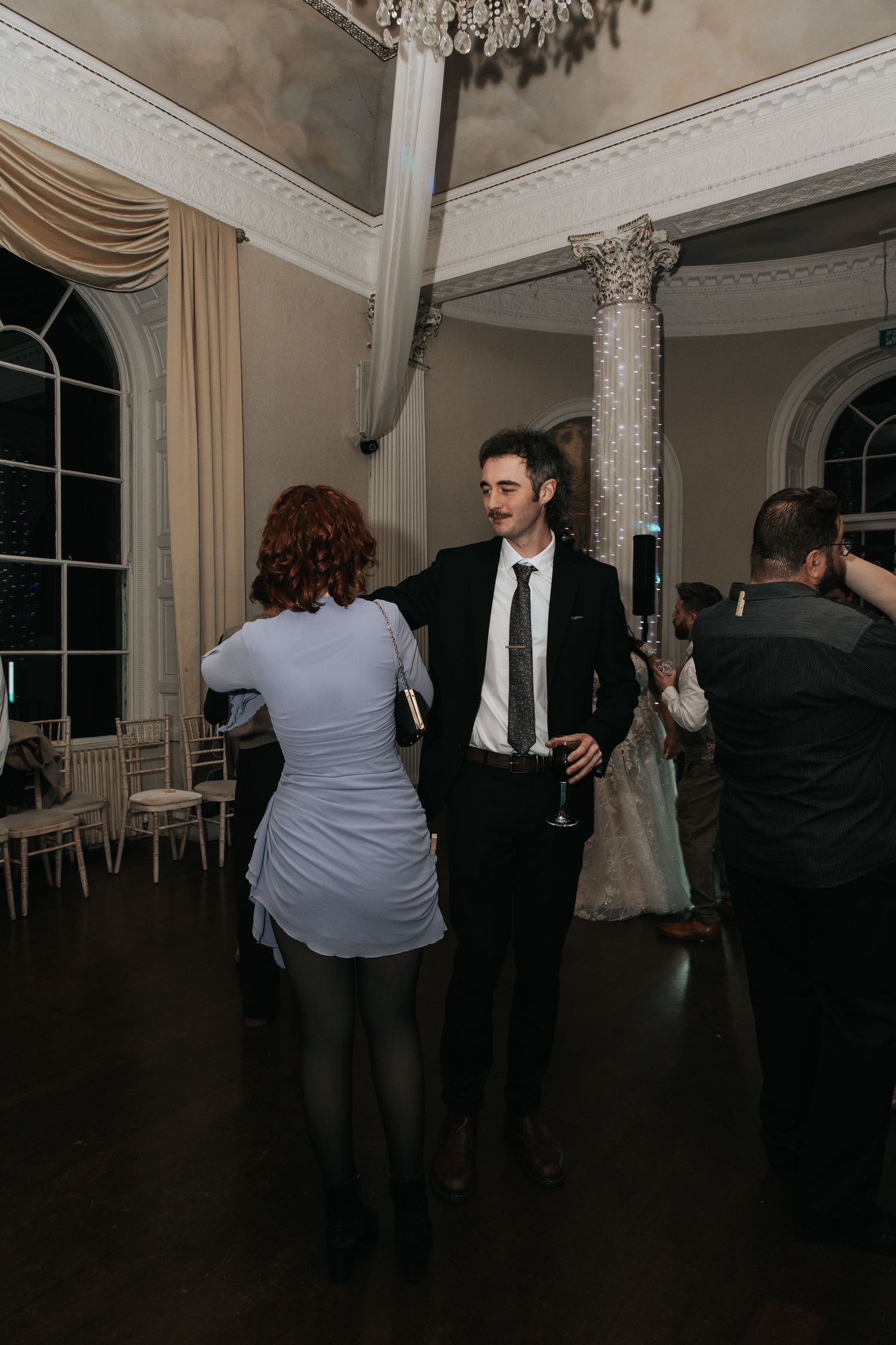 Couples dancing together at an elegant wedding reception in a grand room with ornate columns and chandelier.