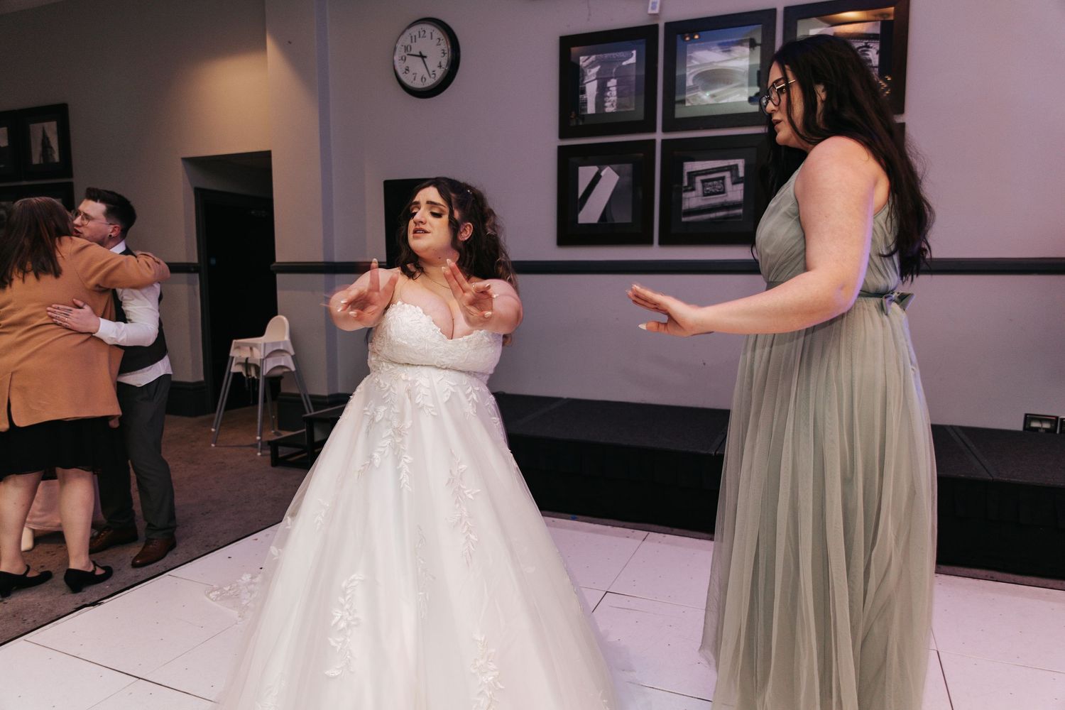 A person in a sparkly white wedding gown dances with a bridesmaid in sage green on a white dance floor.