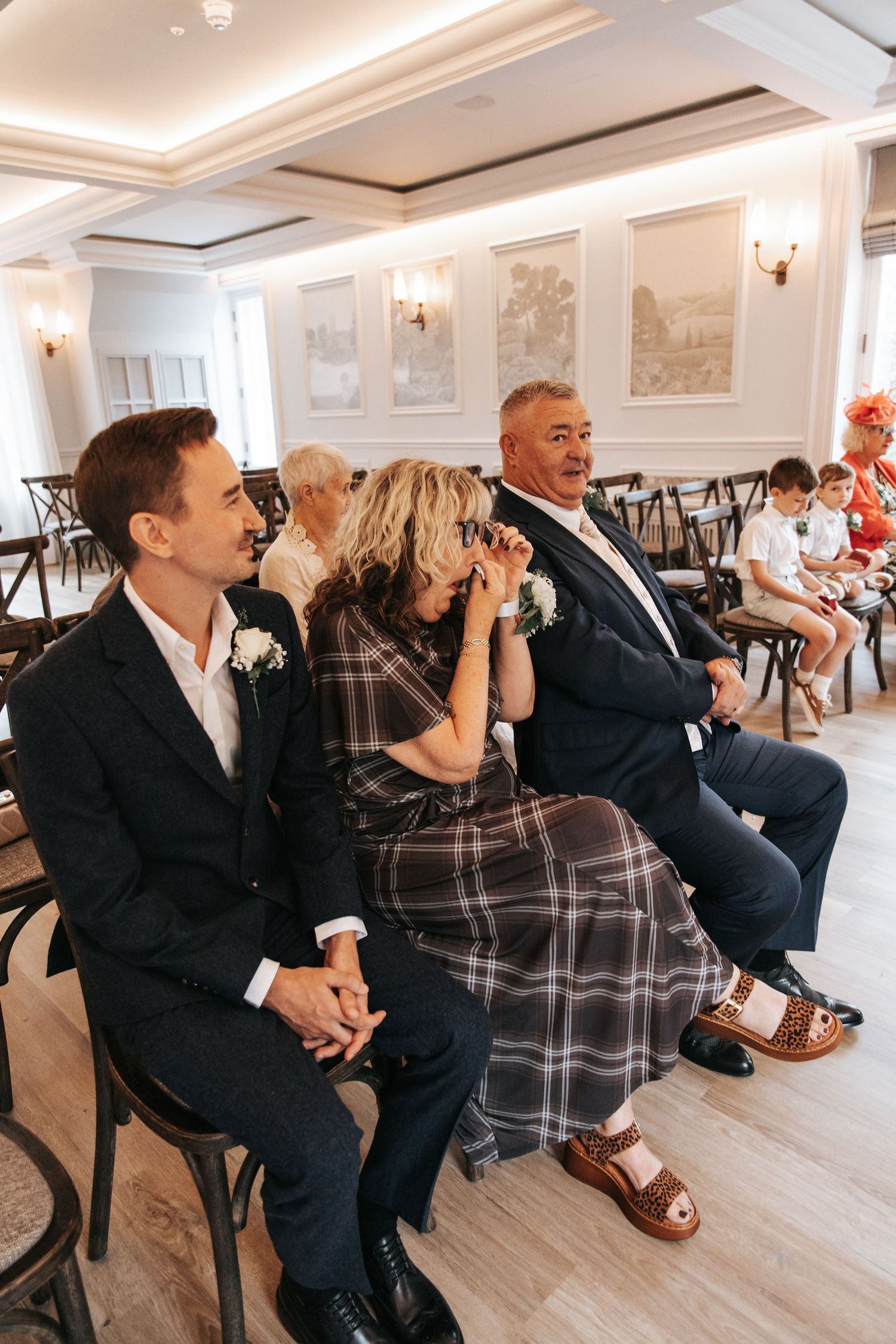 Emotional wedding guests seated in elegant venue, woman wiping tears, men in suits with boutonnieres.