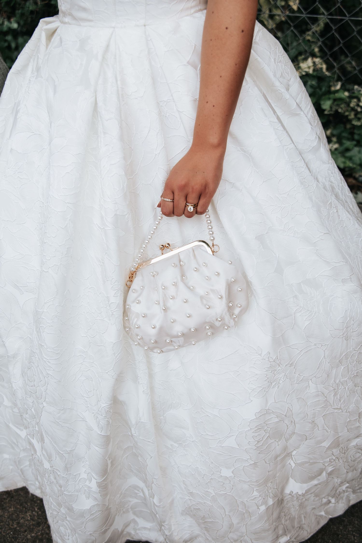 Bride in white lace gown holding elegant pearl-embellished clutch purse with gold chain handle and ring detail.