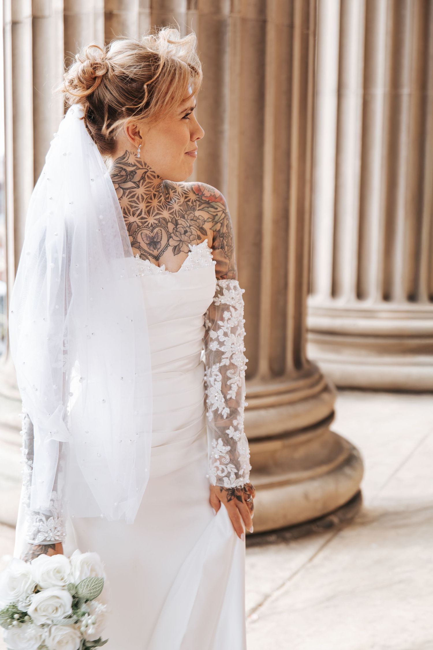 Tattooed bride in white lace wedding dress and veil holding bouquet beside stone columns.