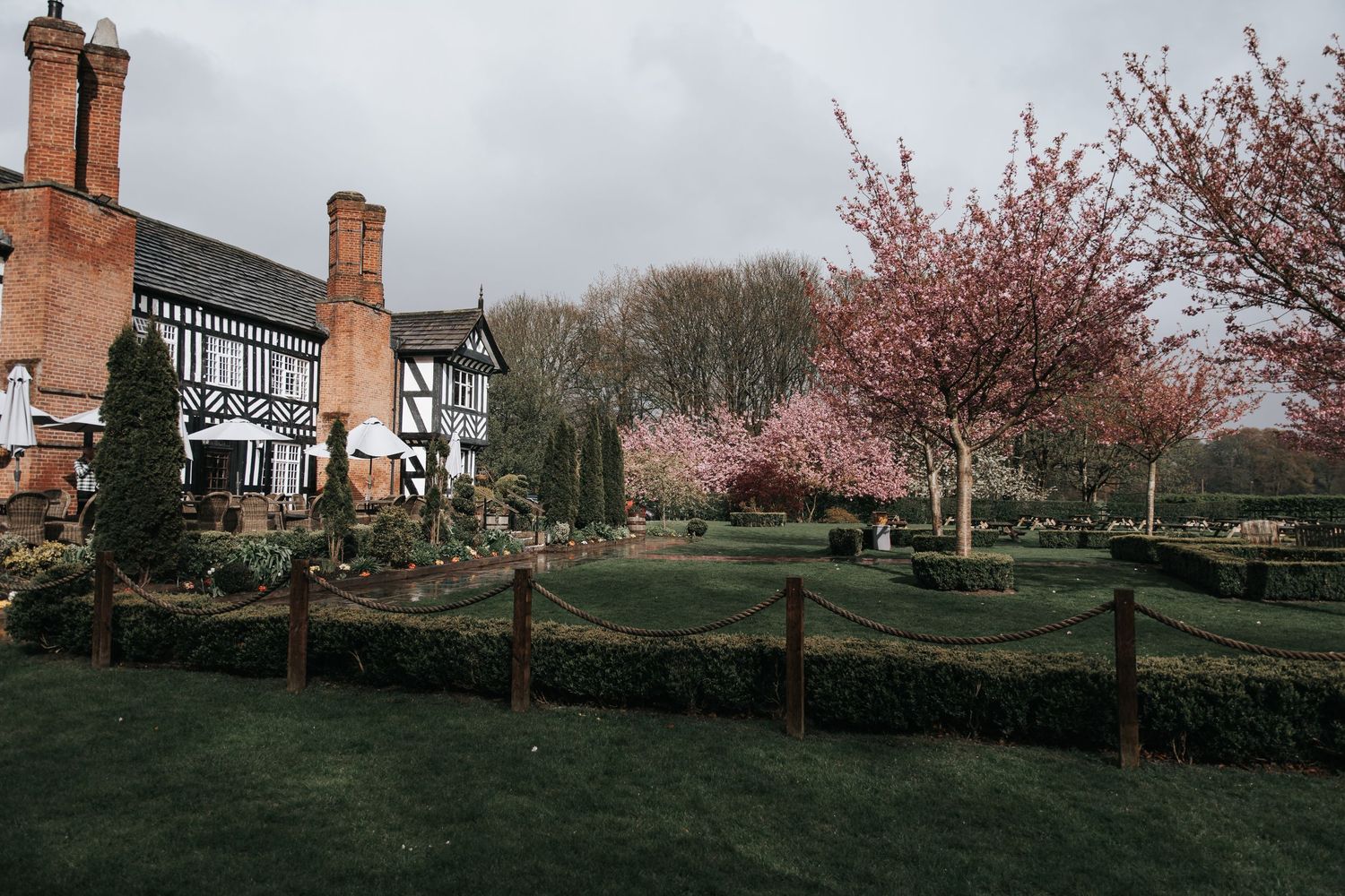Tudor-style manor house with manicured gardens, rope fencing, and pink cherry blossom trees under a cloudy spring sky.