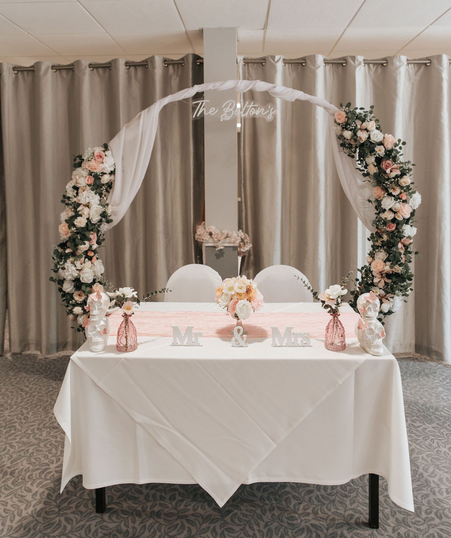 Elegant wedding sweetheart table with floral arch, white draping, pink roses, and Mr. & Mrs. signs in a banquet hall.