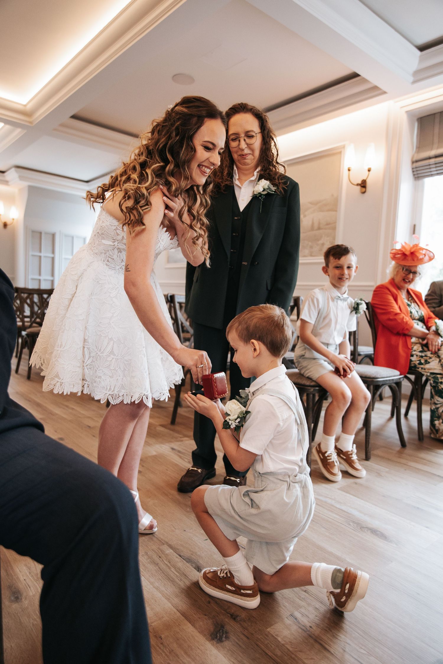 Bride in white lace dress shares a joyful moment with a young ring bearer during an elegant indoor wedding ceremony.